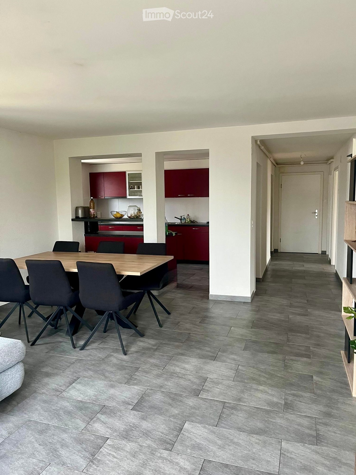 Open-plan living and dining area with grey tiled flooring, a wooden dining table surrounded by black chairs, and a modern kitchen with red cabinets and stainless steel appliances.
