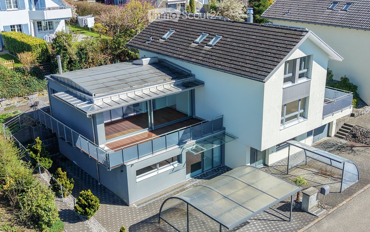 Modern two-story house with a flat roof, large windows, and a spacious balcony area. The house features a sleek design with a combination of light-colored walls and dark roofing. The surrounding area includes a paved driveway, small garden, and neighborin
