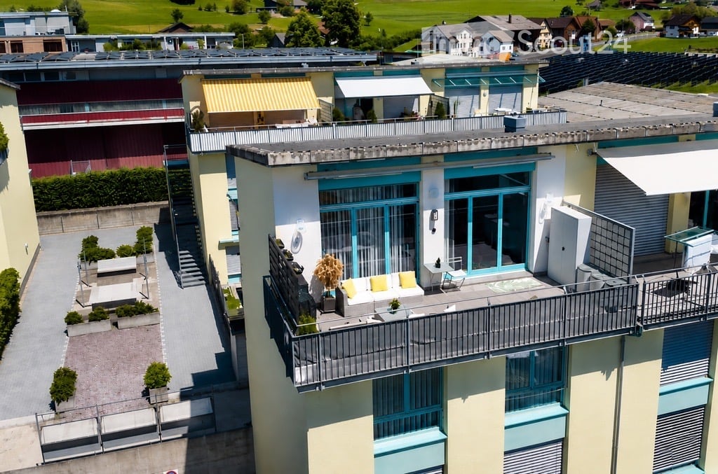 A modern multi-story building with balconies and terraces, featuring outdoor seating areas, potted plants, and a view of a green landscape with houses in the background.