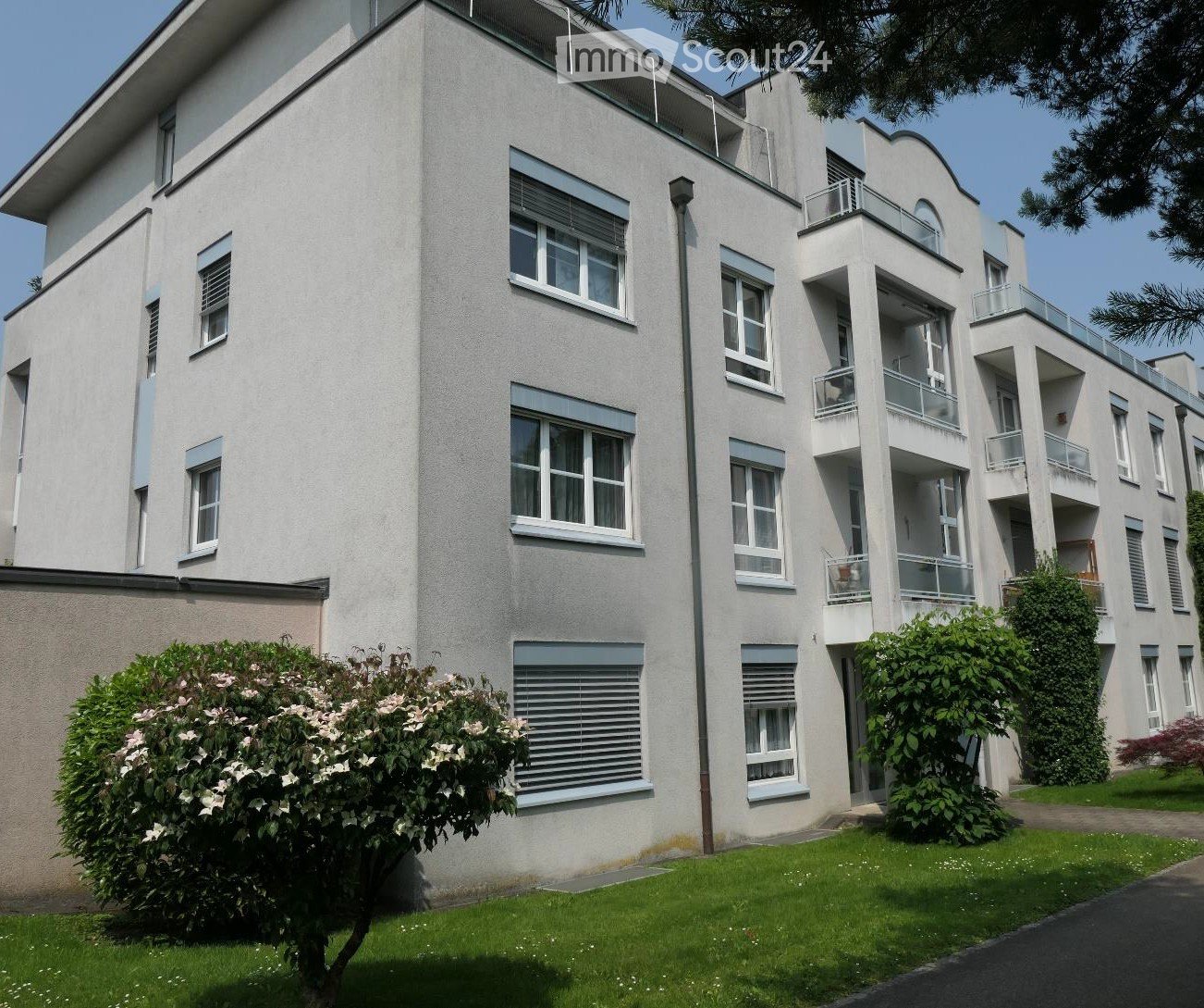 Modern multi-story residential building with white facade, multiple windows, balconies, and surrounding greenery.