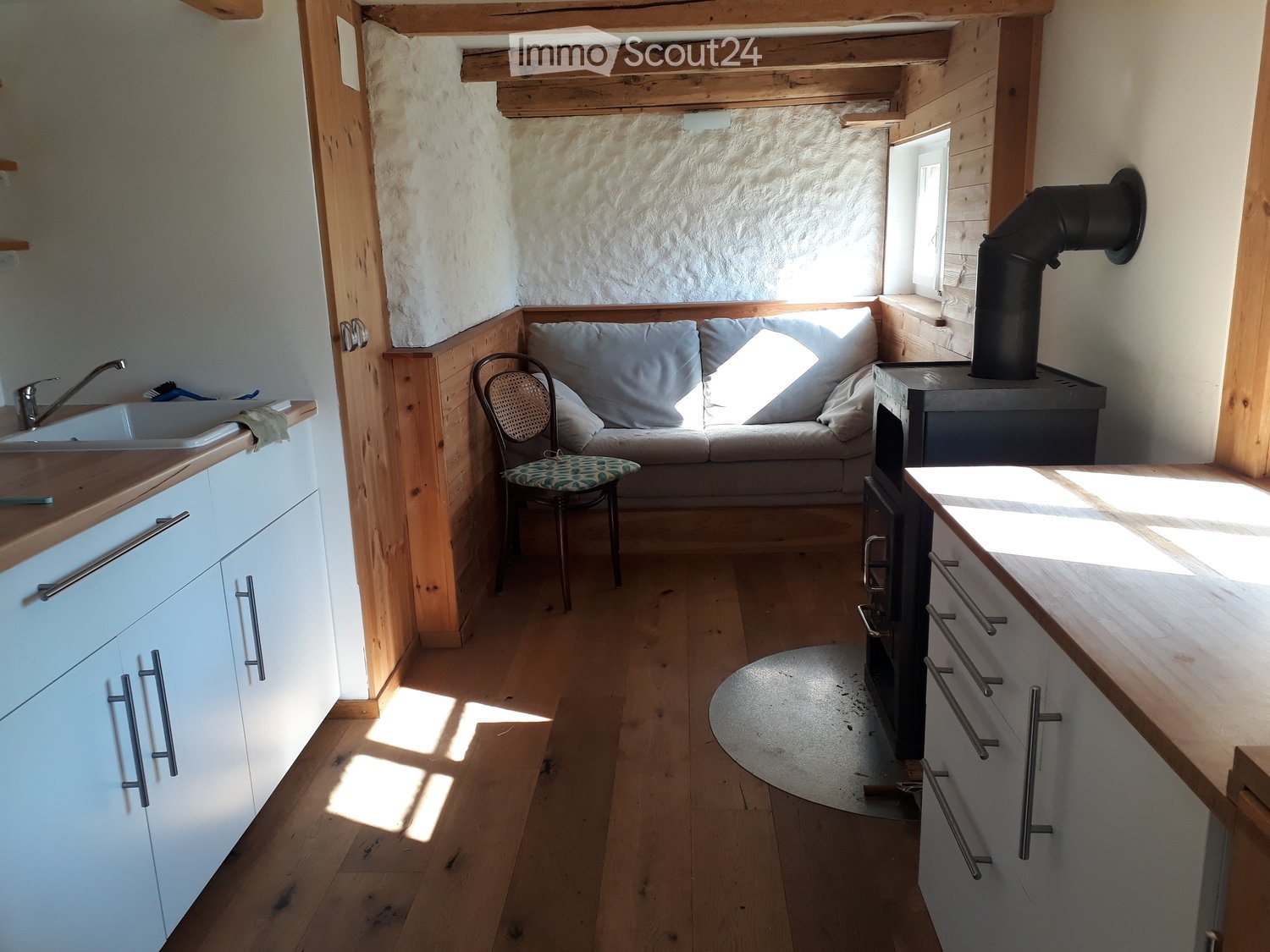 Cozy, compact living space with a sofa, chair, wood stove, sink, and cabinets, featuring wooden floors and natural light from a window.