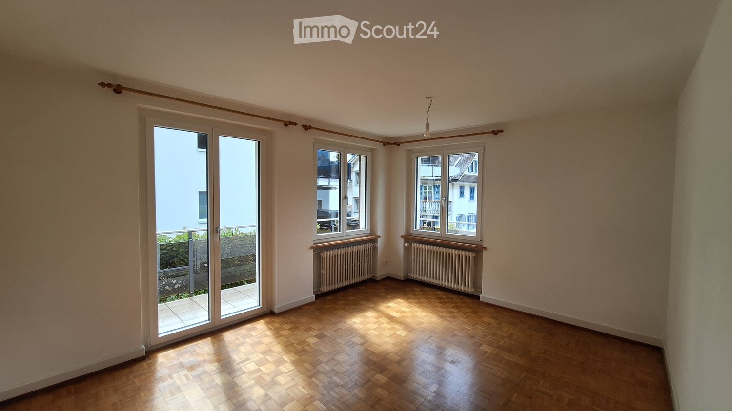 Empty room with wooden flooring, large windows, radiators, and a sliding glass door leading to a balcony.