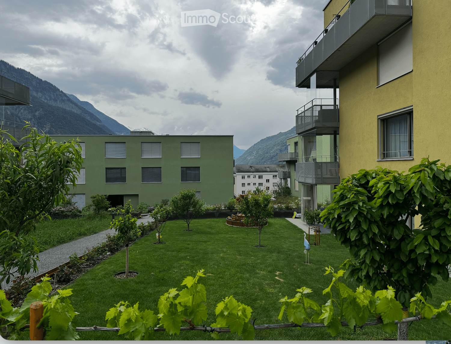 Well-maintained garden area with green grass, small trees, and shrubs, surrounded by modern apartment buildings and a scenic mountain backdrop.