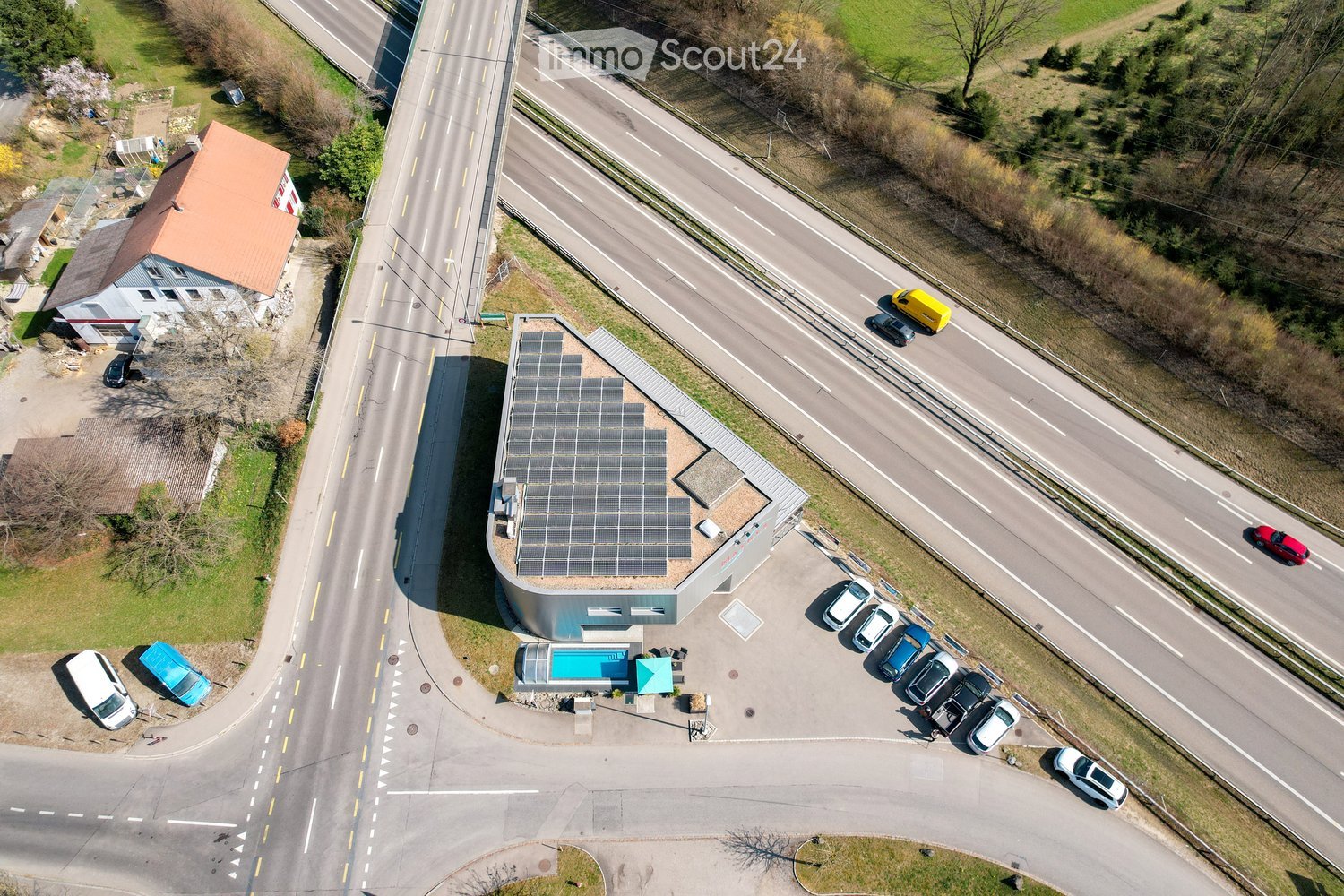 Aerial view of a modern building with a solar panel roof, surrounded by a parking lot with several cars, a small pool, and a nearby road with traffic.