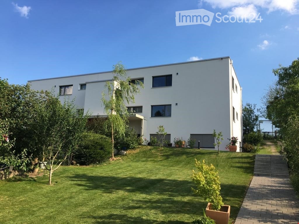 Two-story house with modern architectural design, featuring a white exterior, large windows, and a balcony on the second floor. The property is situated on a sloping terrain with a driveway leading to the entrance. The garden includes lush greenery, shrub