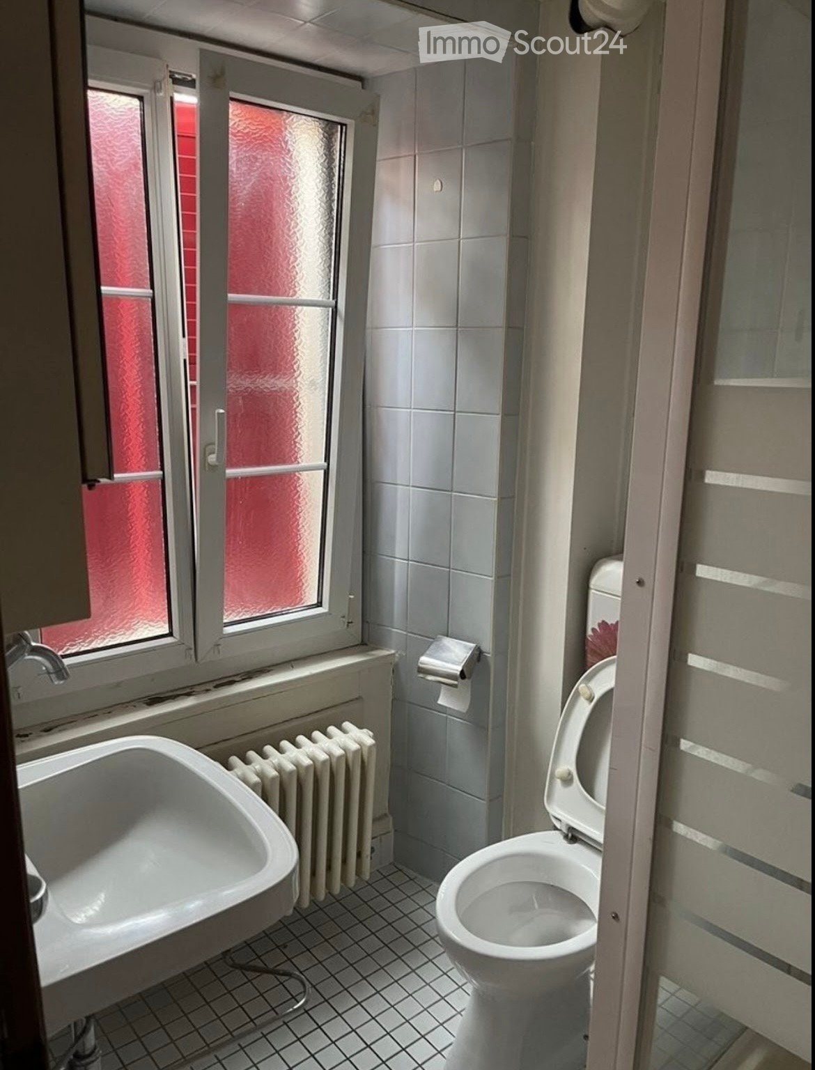 Bathroom with a toilet, sink, radiator, and toilet paper holder. The room has tiled walls and floor, and a window with frosted glass.