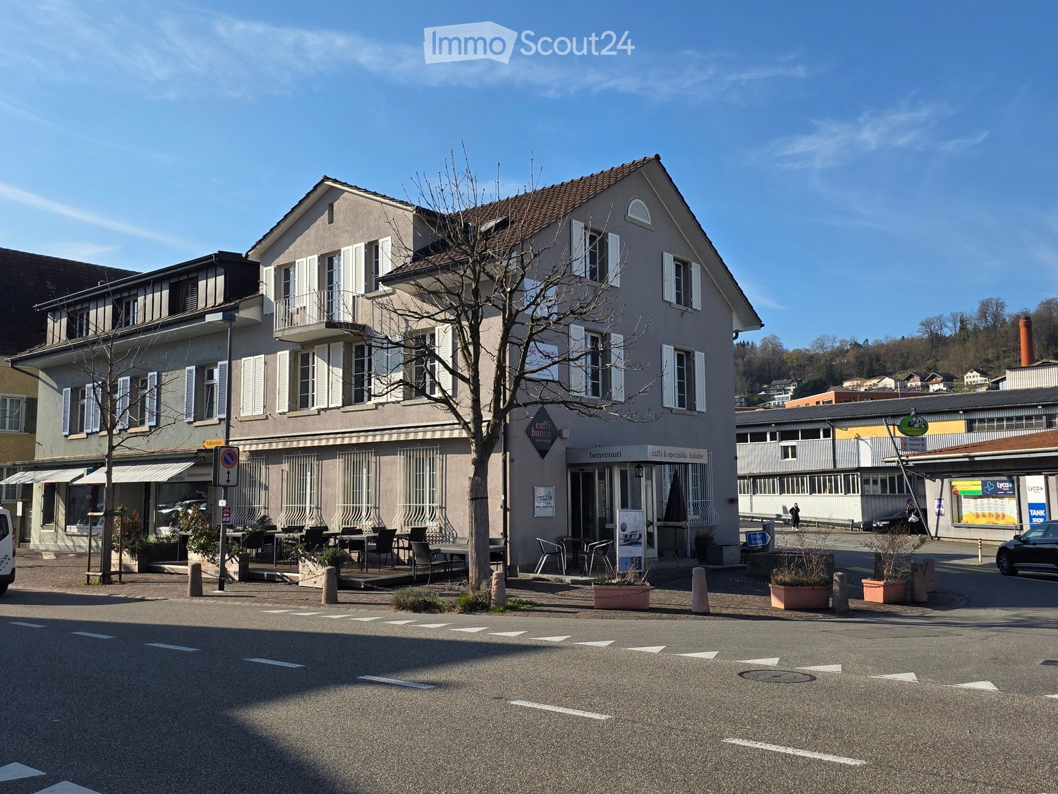 Two-story building with a cafe on the ground floor, outdoor seating area, and multiple windows with shutters. The building has a light-colored facade and is situated on a street corner with a tree in front.