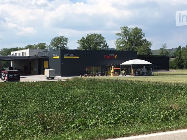 Modern commercial building with a black facade, signage for "Neff AG" and "Tenne Grill", outdoor seating area, and a grassy lawn in front.