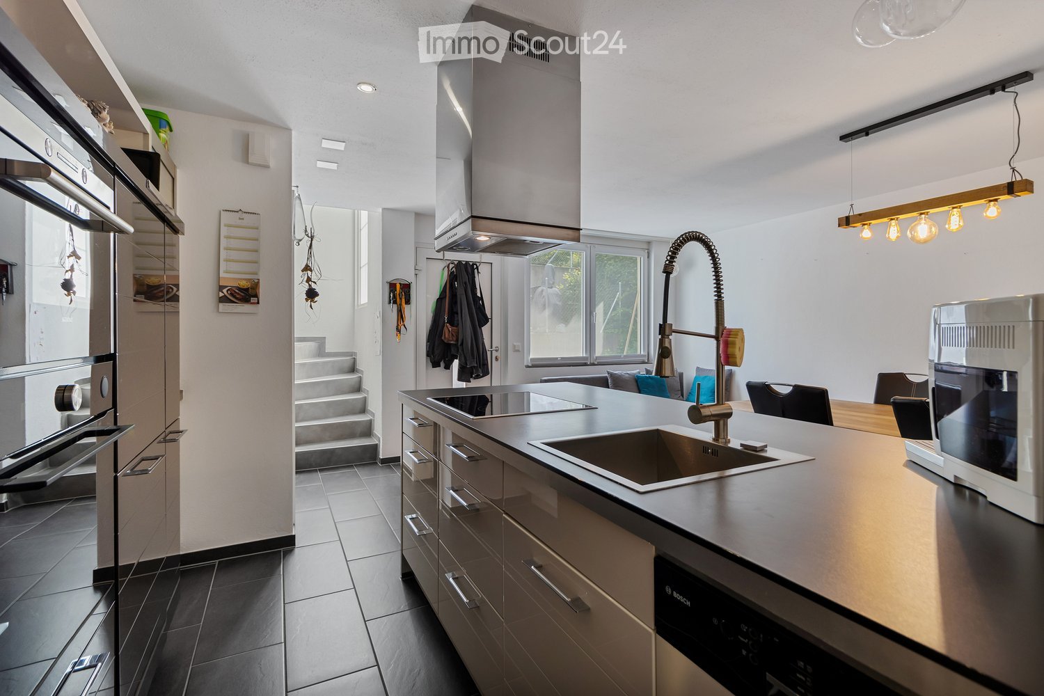 Modern open kitchen with stainless steel appliances, central island, black countertops, gas stove, silver chimney hood, sink, faucet, dishwasher, white cabinets, tiled flooring.