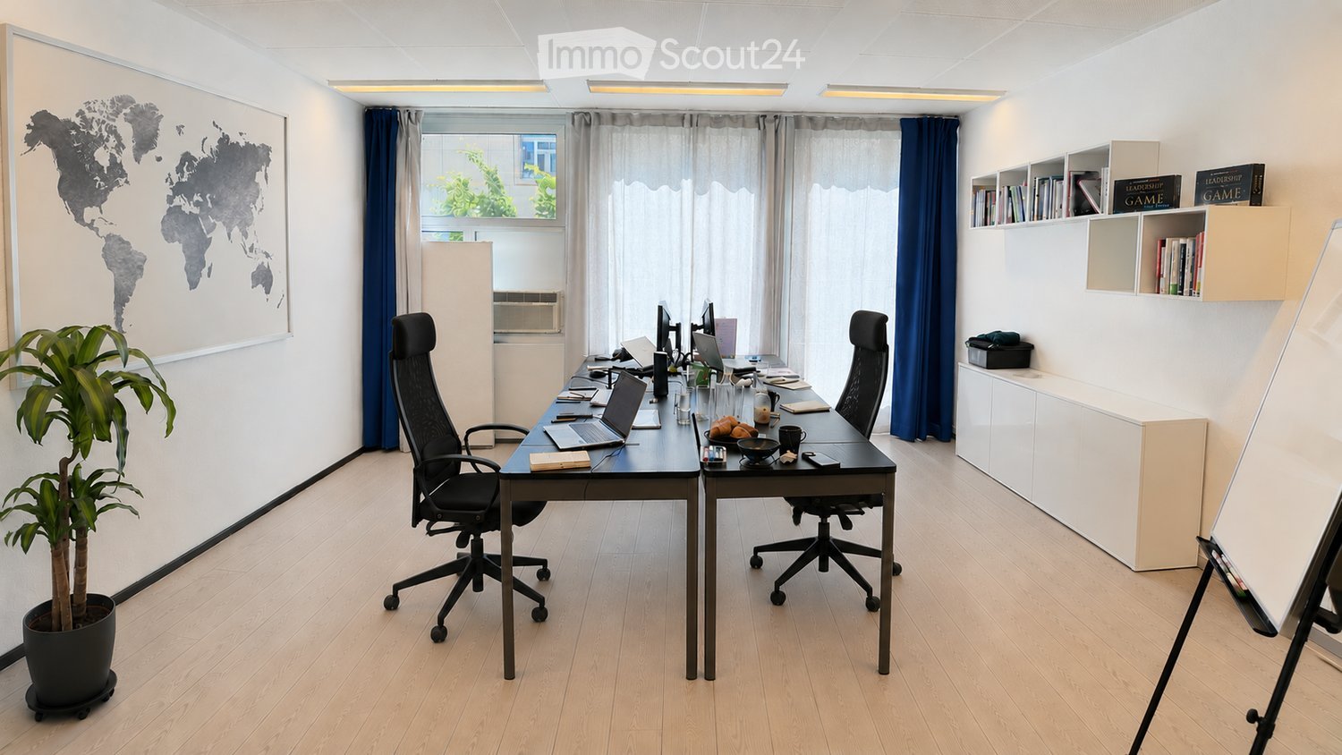 Modern office space with a large desk, two ergonomic chairs, multiple laptops, screens, books, and a potted plant. Shelves with books and decorative items are visible.