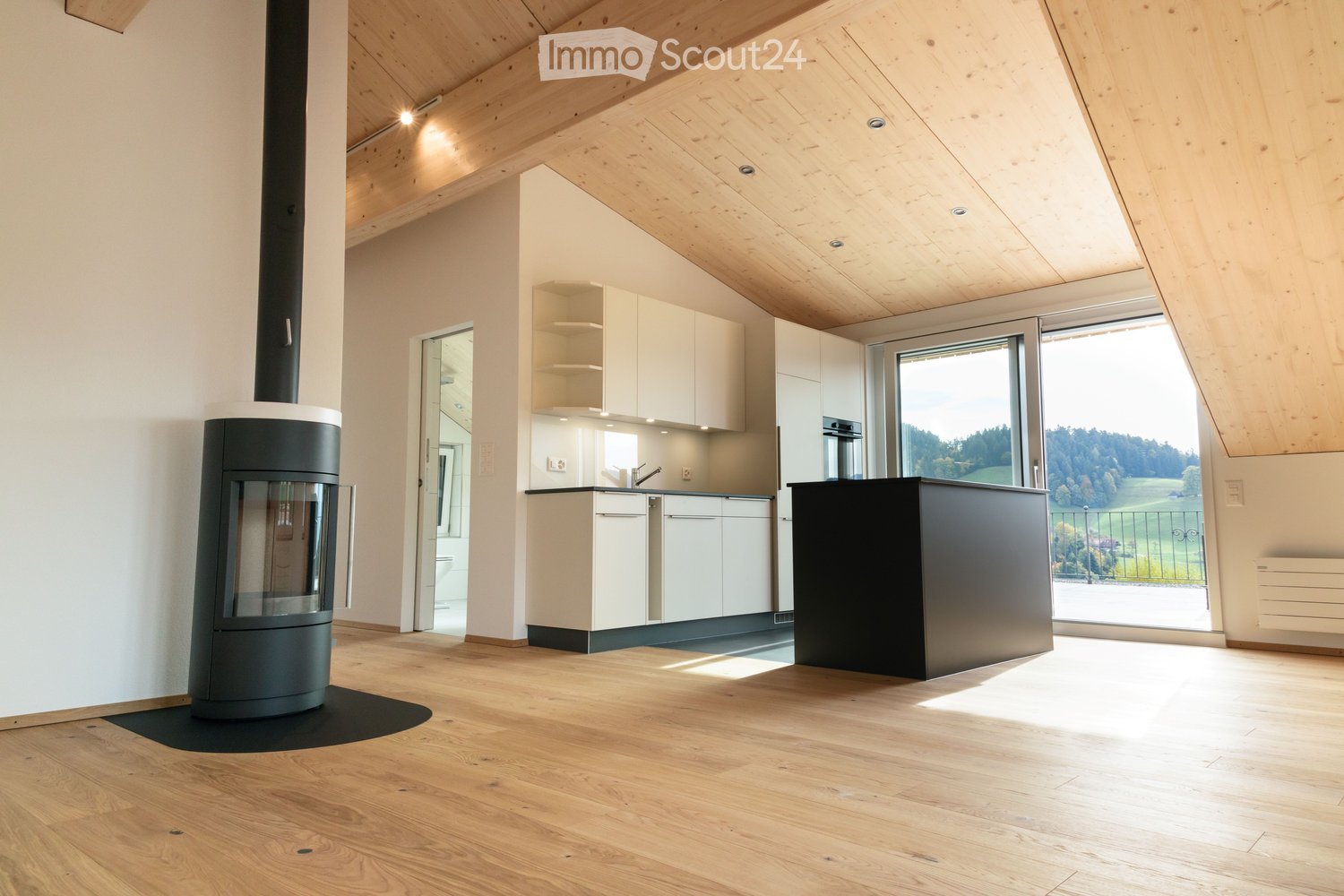 Open-plan living and kitchen area with wooden flooring, white cabinets, a central island, and a modern fireplace. Large windows offer a scenic view of the landscape.
