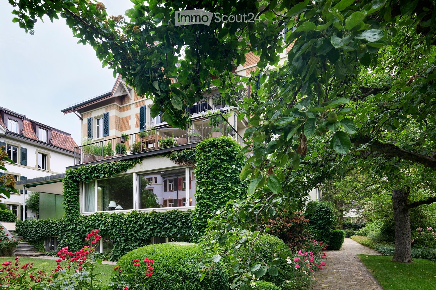 A lush garden area with well-maintained hedges, flowering bushes, and a winding brick pathway. The garden is surrounded by residential buildings with ivy-covered walls and balconies.