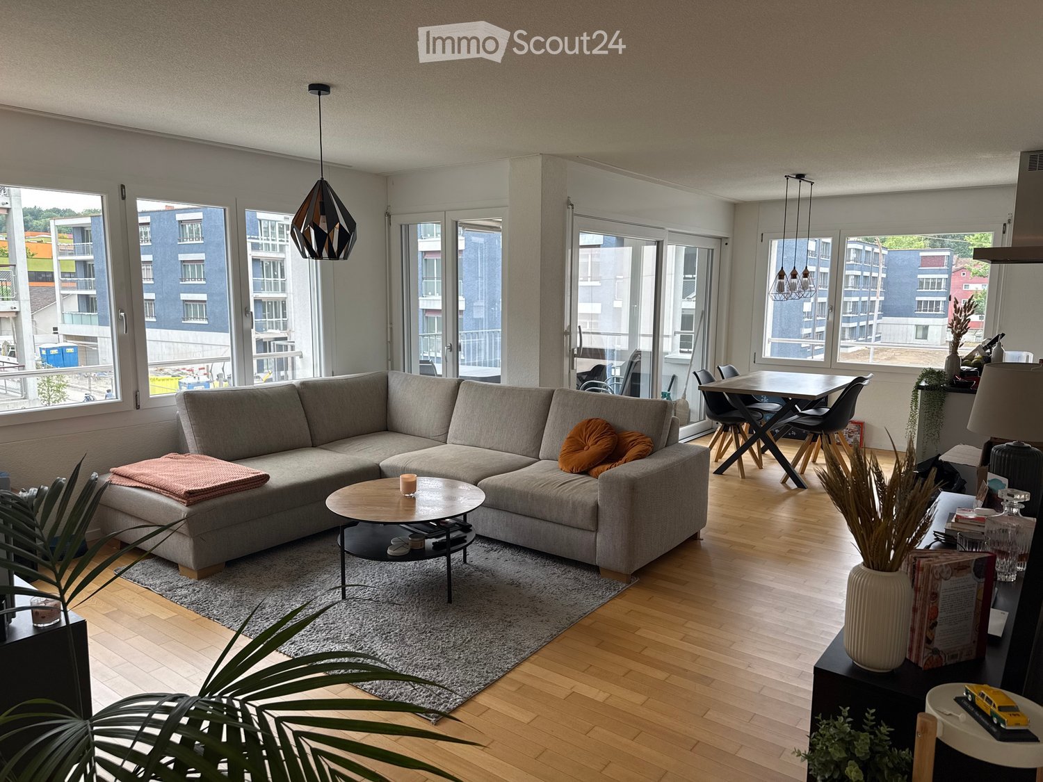 Modern living room with large windows, grey sectional sofa, coffee table, side table, dining area with chairs and table, decorative plants, and lamps.
