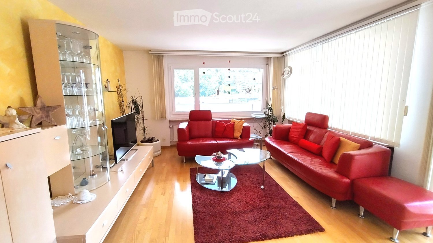 Modern living room with red leather sofas, a glass coffee table, a TV stand, a glass cabinet, and a plant near the window.