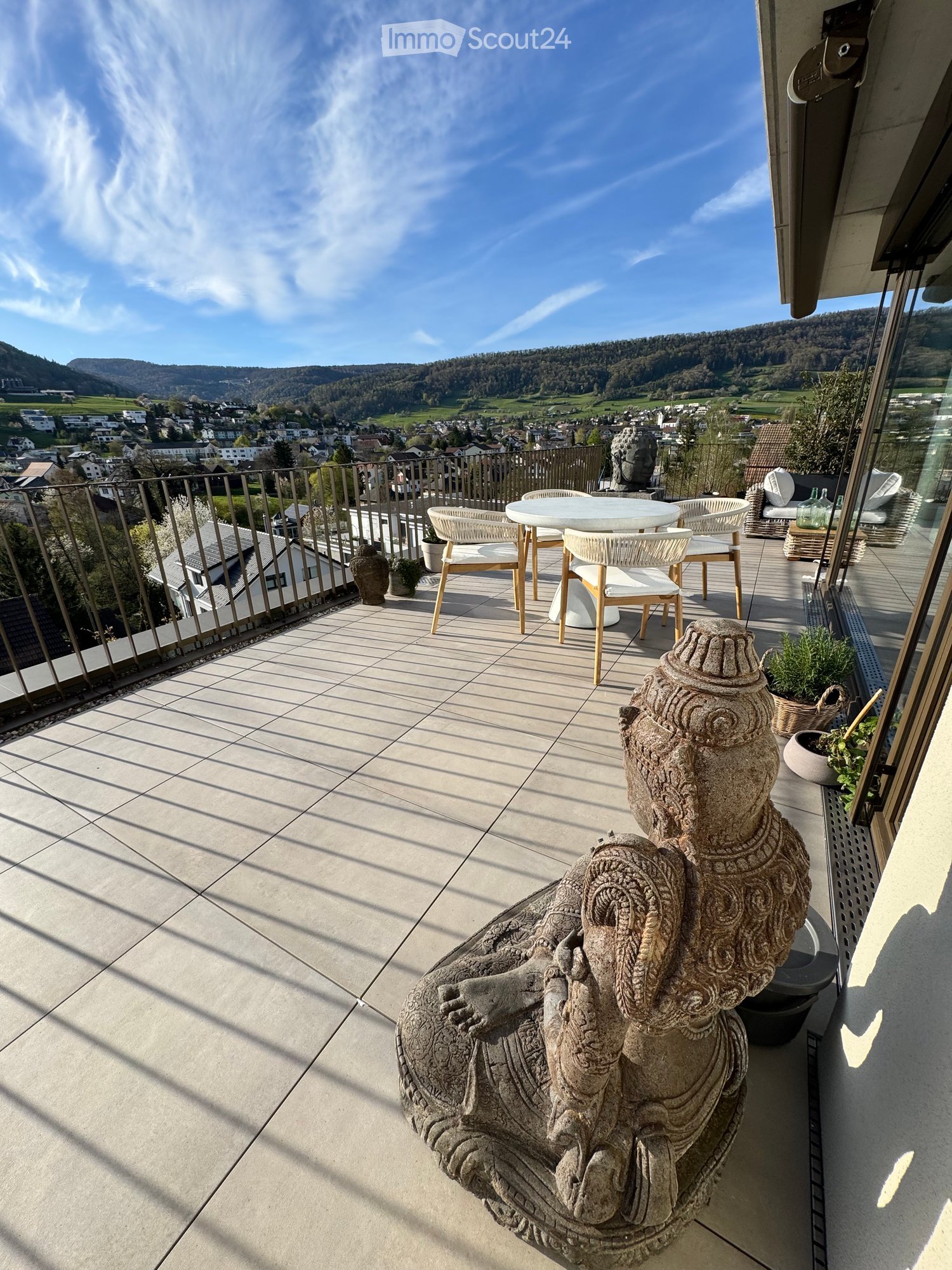 A balcony with a table and chairs, a statue, and a view of the mountains and town