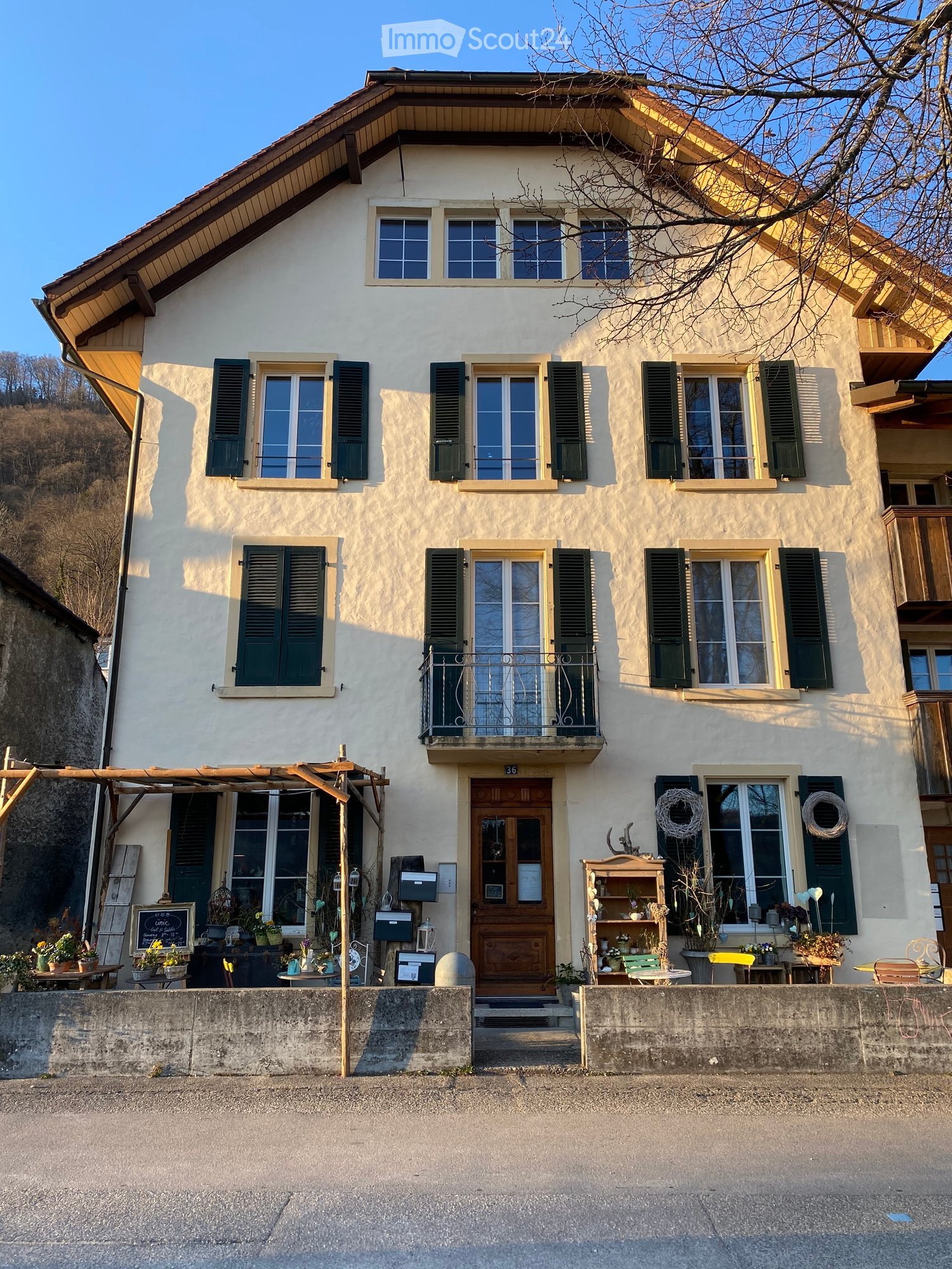 Two-story house with light-colored walls, green shutters, and a balcony on the second floor. The entrance features a wooden door and a small porch area with decorative items and plants.