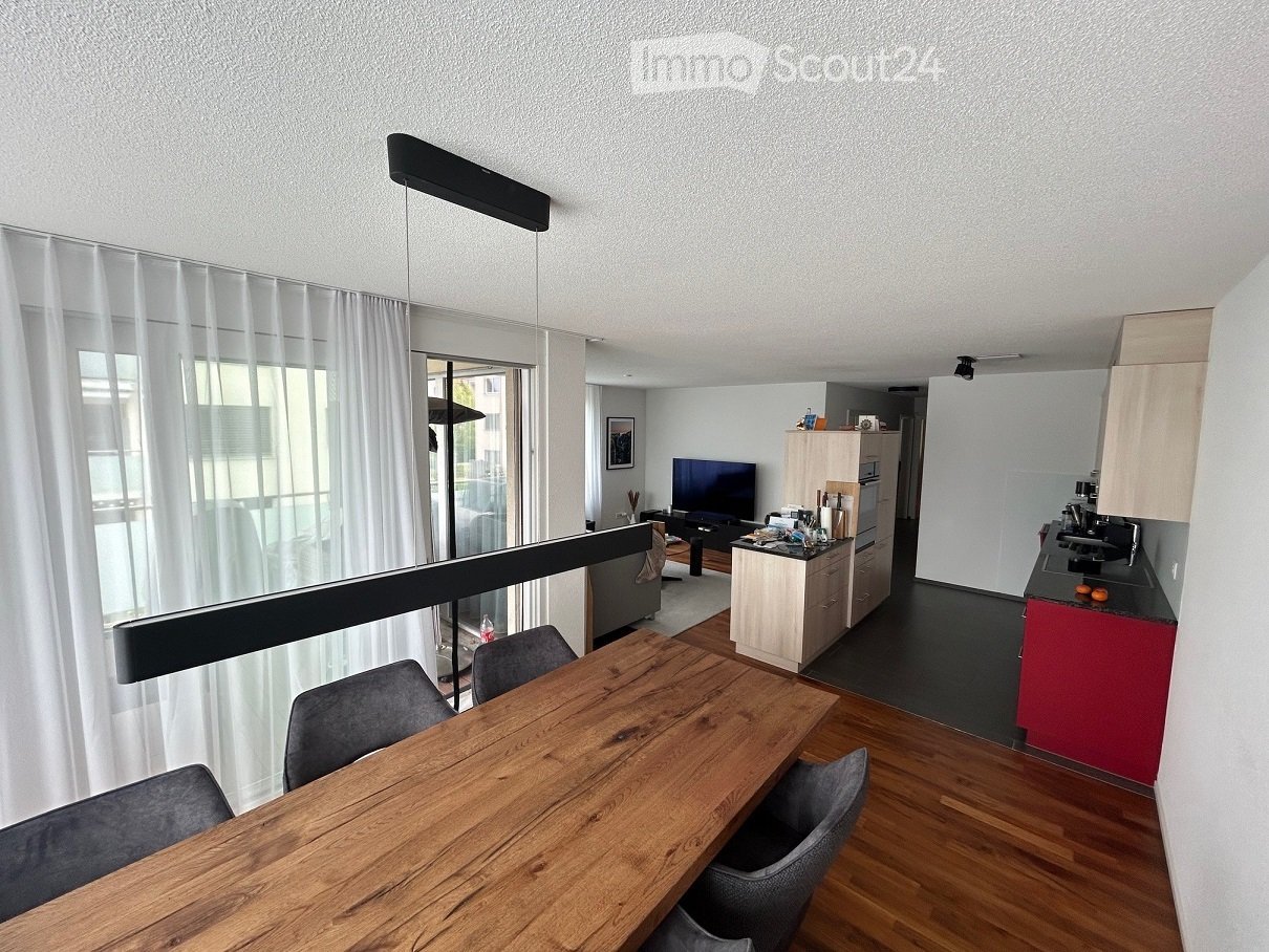 Open-plan living and dining area with a wooden dining table, chairs, a television, a small kitchen area with a sink, refrigerator, and a balcony with sheer curtains.