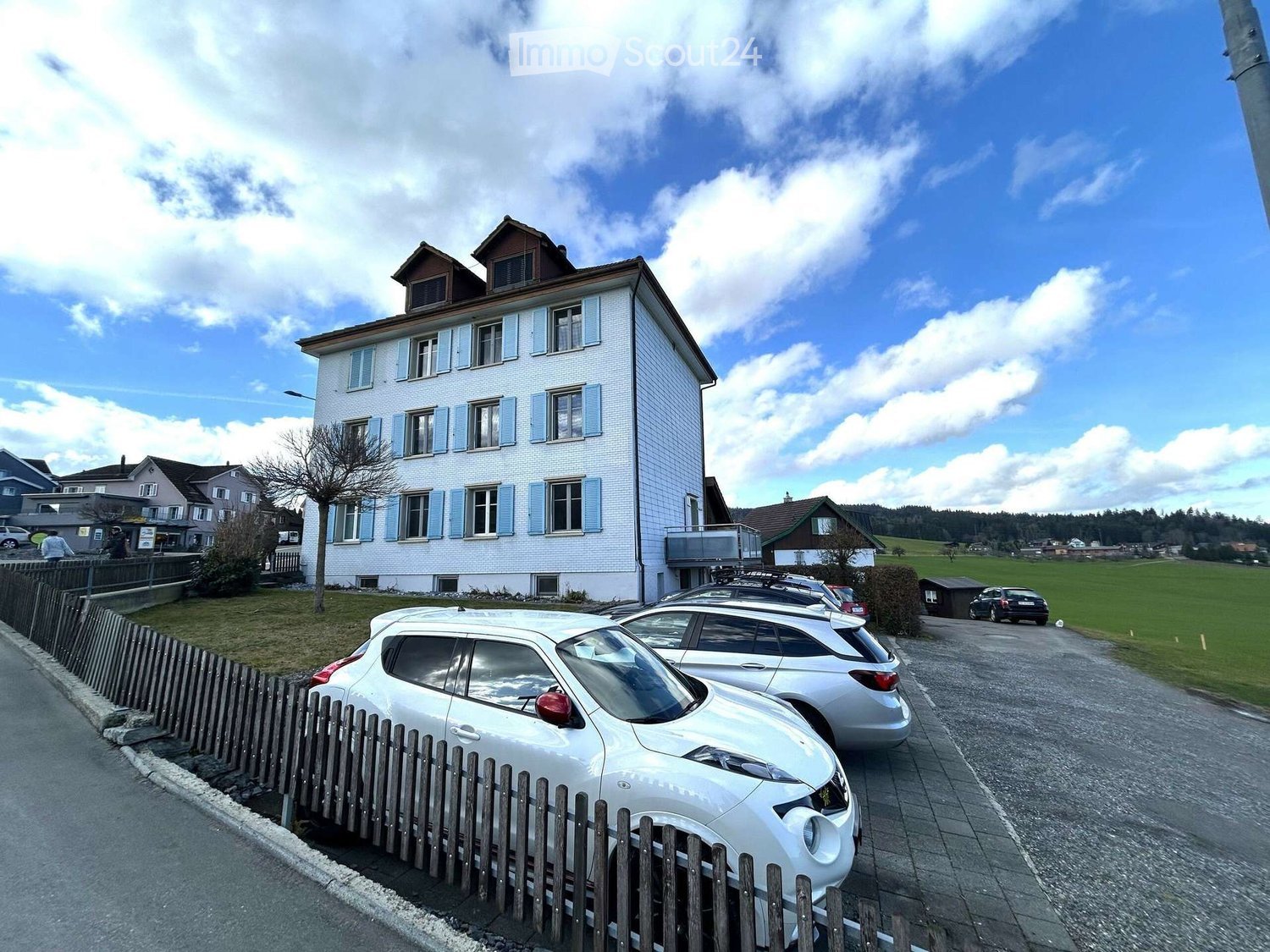 A two-story white house with blue shutters, multiple windows, and a small balcony on the right side. Several cars are parked in front of the house, and there is a wooden fence along the sidewalk.