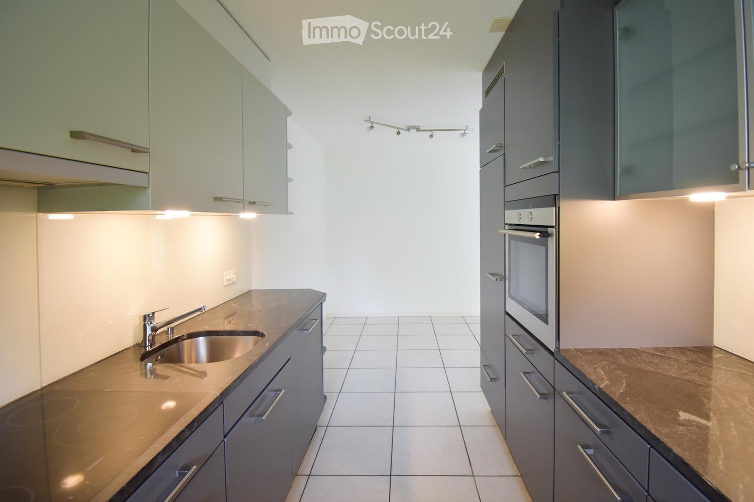 Modern kitchen with sleek grey cabinets, stainless steel sink, and built-in oven. The countertops are light-colored, and the space is well-lit with integrated lighting.