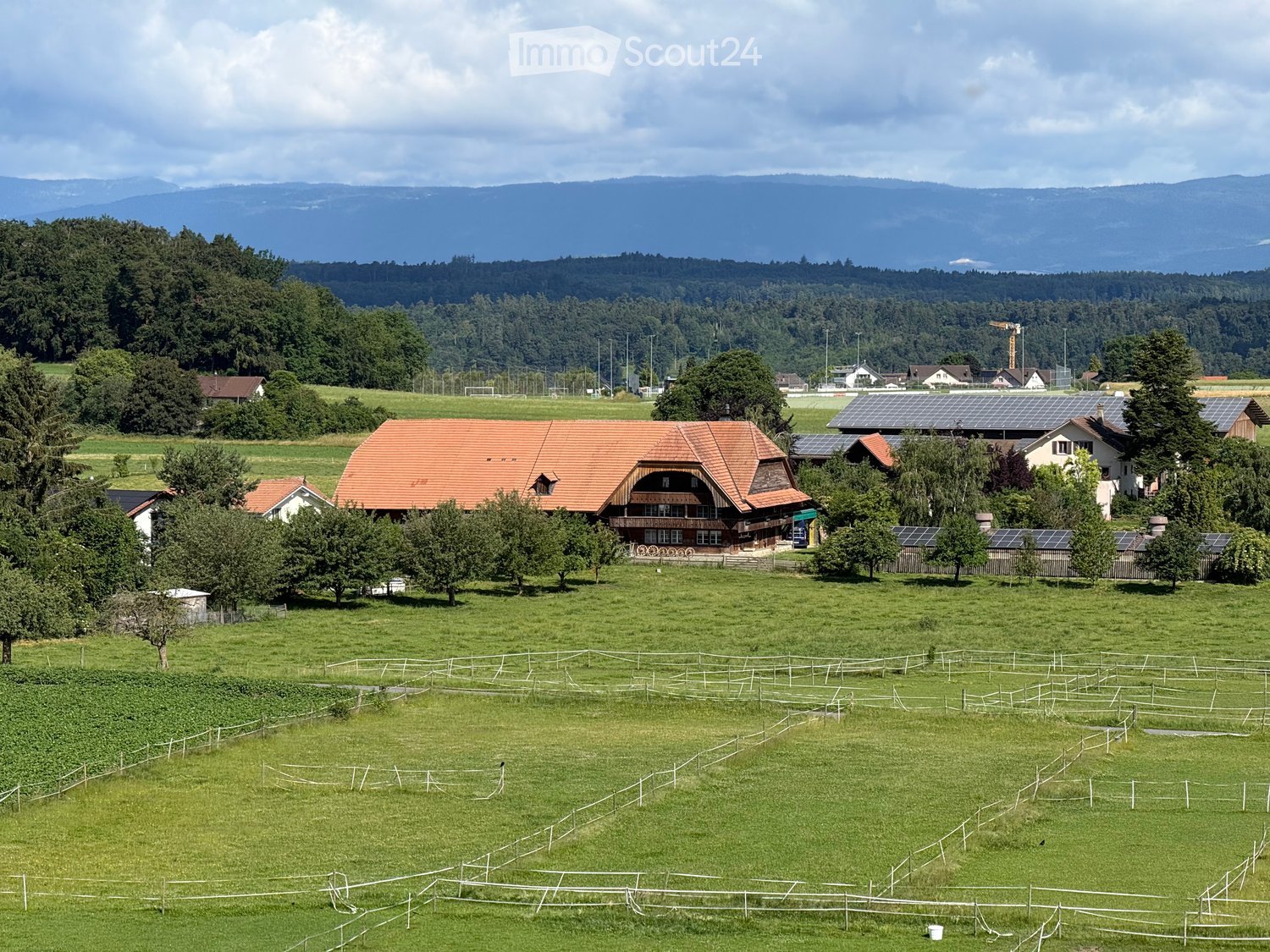 A rural landscape with a large house featuring a red-tiled roof, surrounded by green fields, trees, and solar panels on nearby structures. The area includes fenced enclosures and distant mountains.