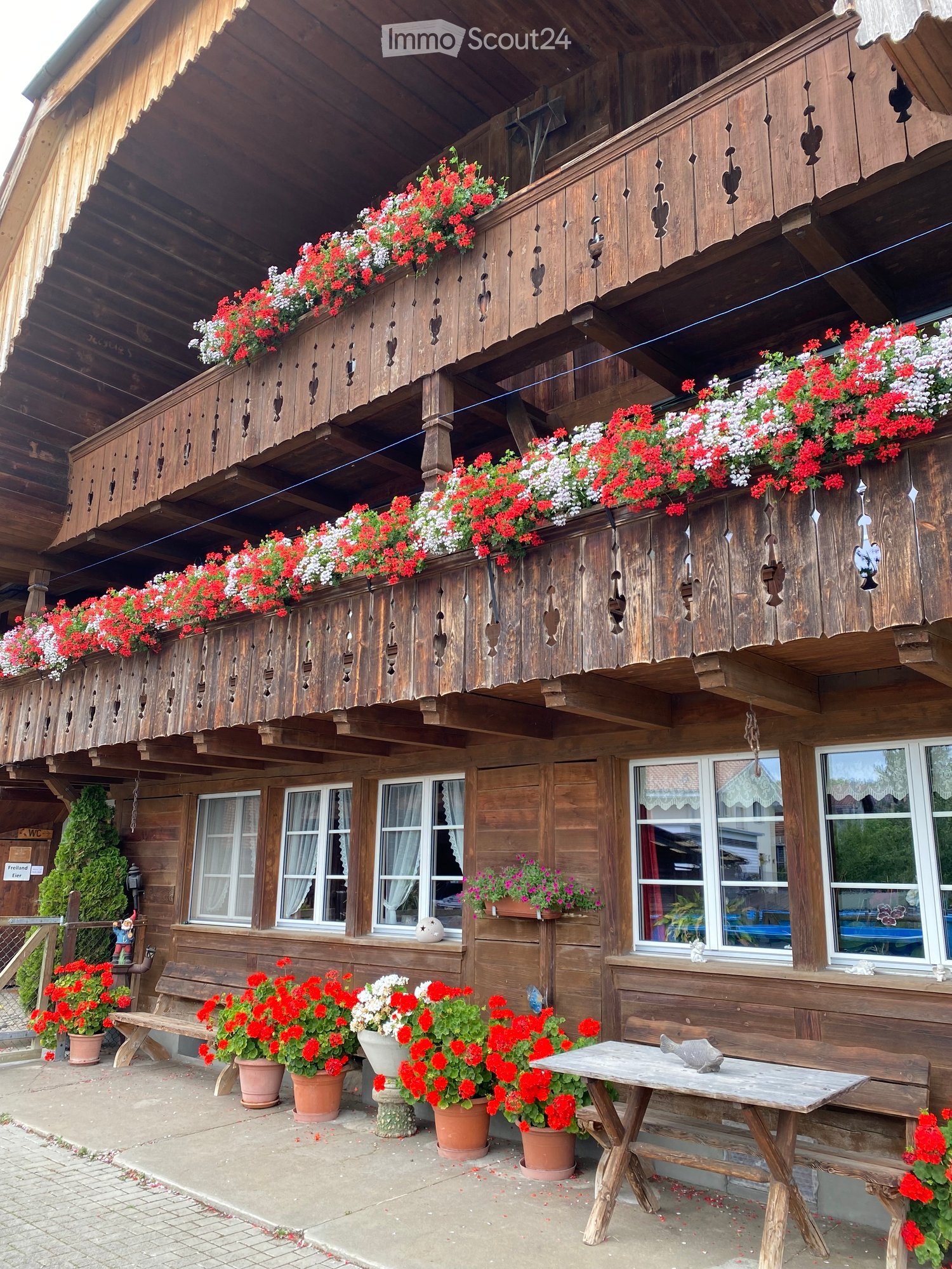 a wooden building with a balcony with flowers, a front bench, a desk, potted plants, white windows, and red flowers in front