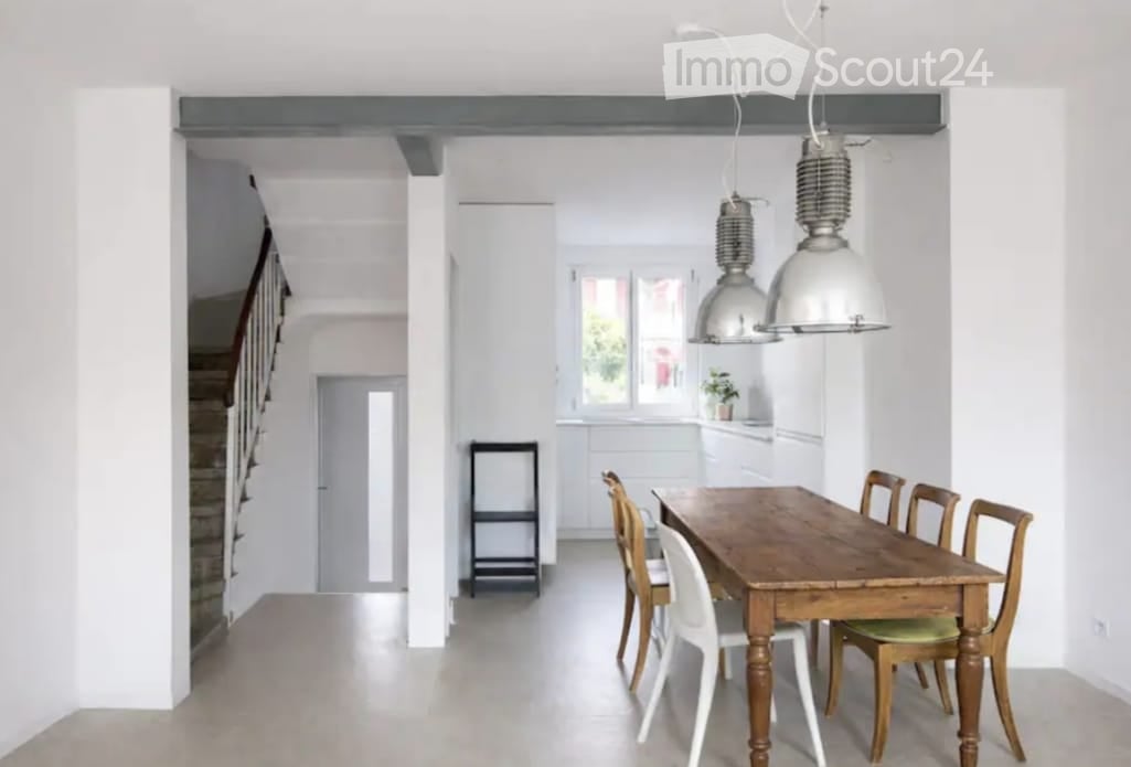 Bright, open-plan dining area with a wooden dining table, white and wooden chairs, pendant lights, and a small stool near the kitchen area.