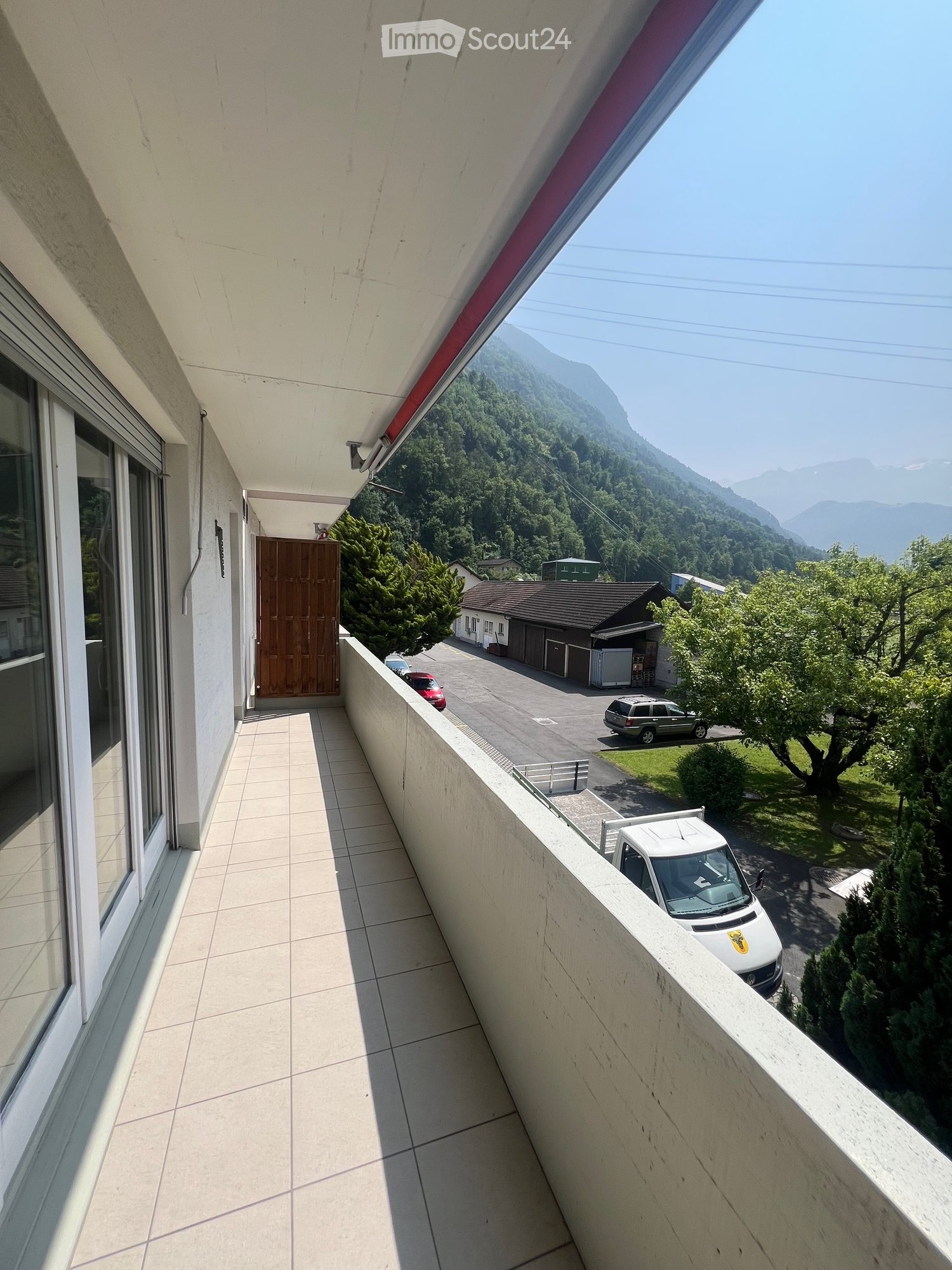 {"visible_features": ["Balcony / Terrace"],"type": "Balcony","description": "The image shows a balcony or terrace with a view of a mountainous, forested landscape. The balcony has a tiled floor and a red awning. There are buildings and a car visible in th