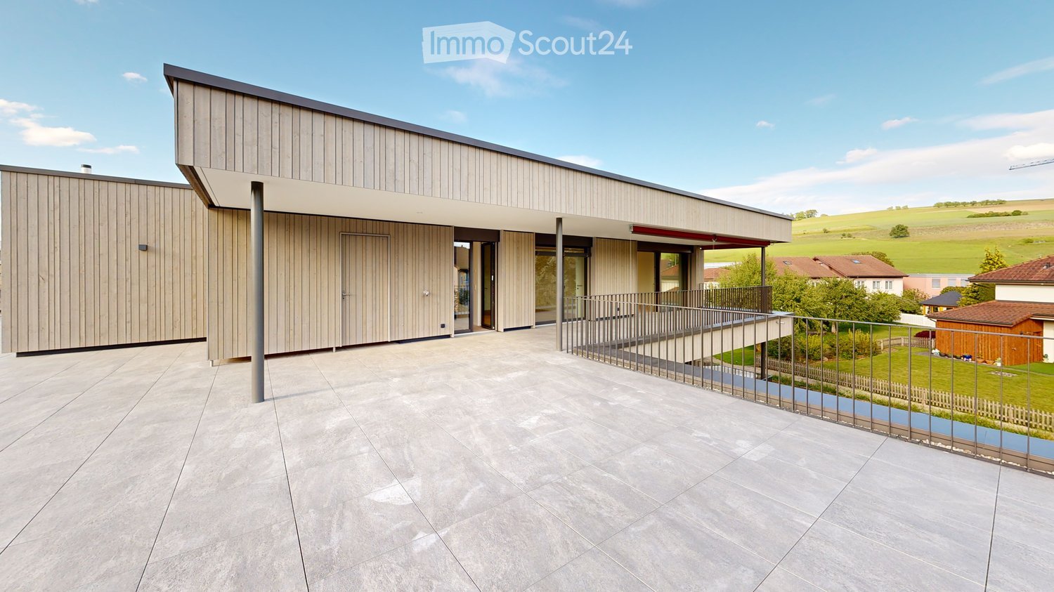 Modern two-story house with a flat roof and wooden facade, featuring a spacious patio with a metal railing and a view of a green landscape.