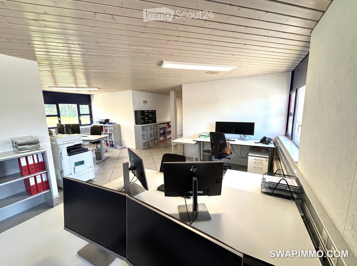 Office space with multiple desks, chairs, computer screens, printers, cabinets, and a telephone.