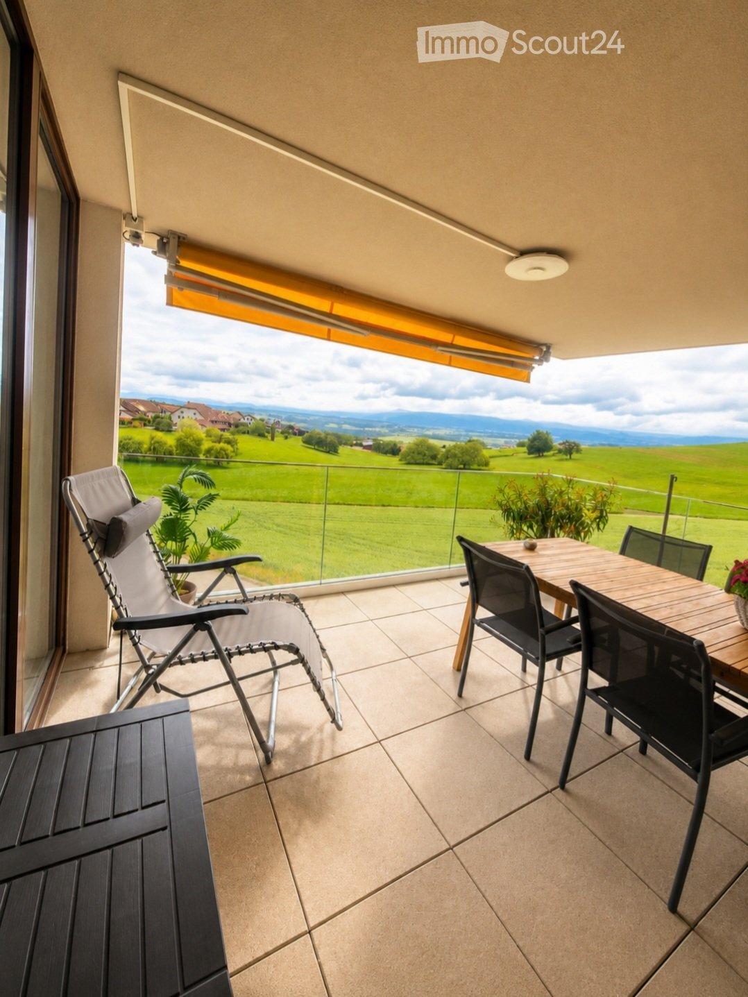 Balcony with tiled floor, glass railing, and scenic countryside view. Includes a wooden table with four black chairs, a folding lounge chair, and potted plants.