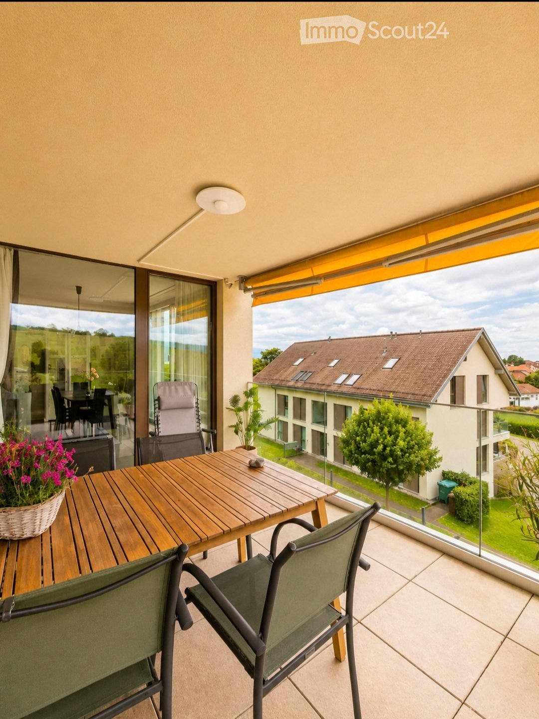 Balcony with wooden table, green chairs, and a flower pot, overlooking a residential area with houses and greenery.