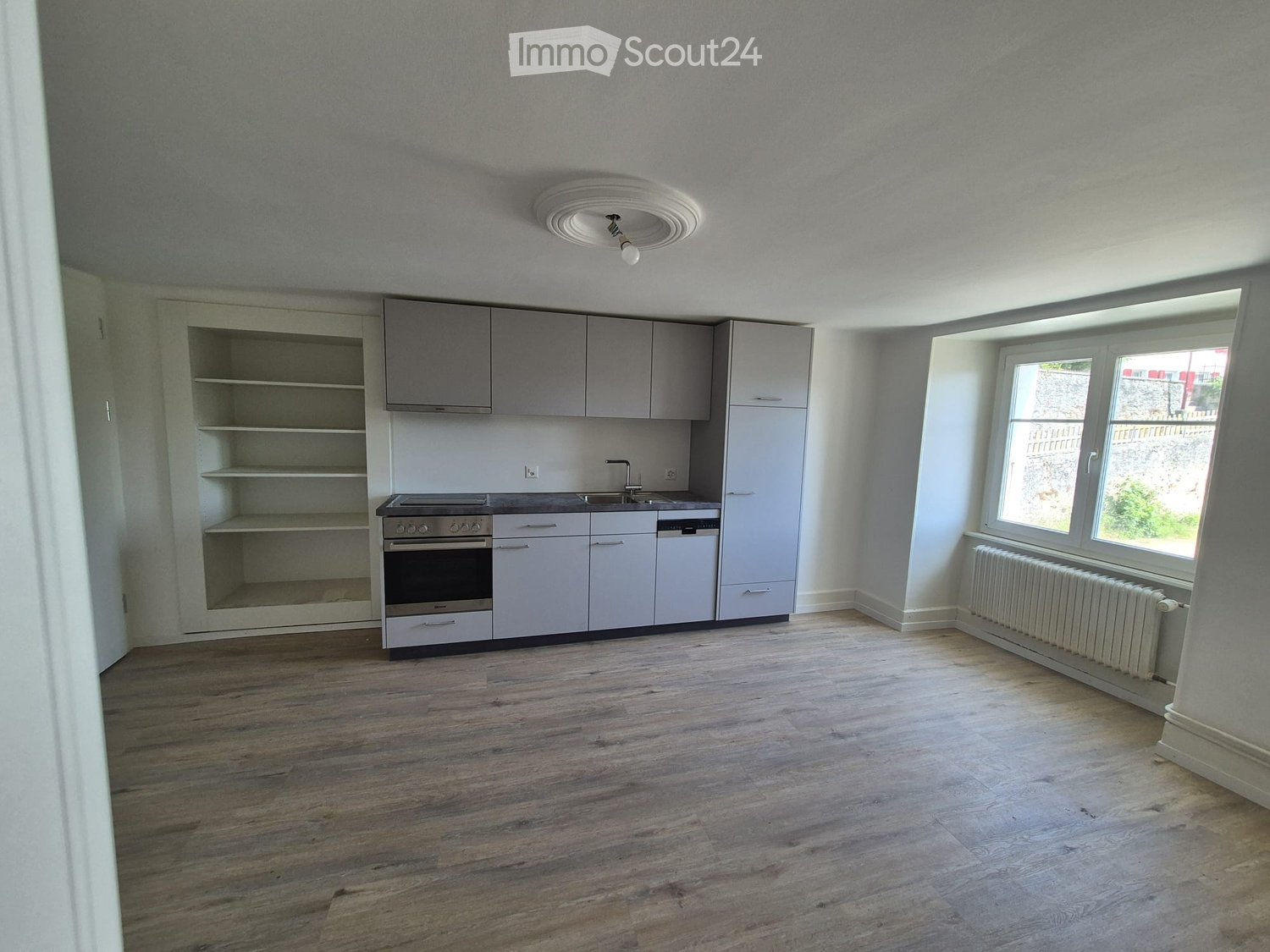 Empty kitchen with light grey cabinets, a built-in oven, sink, and radiator under a large window.