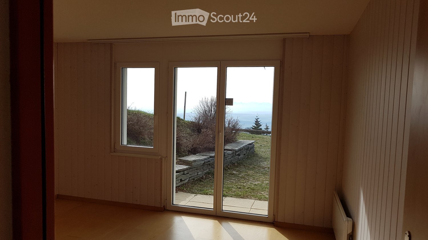 Empty room with wooden walls and floor, large sliding glass doors leading to a grassy outdoor area with a view of the sea.