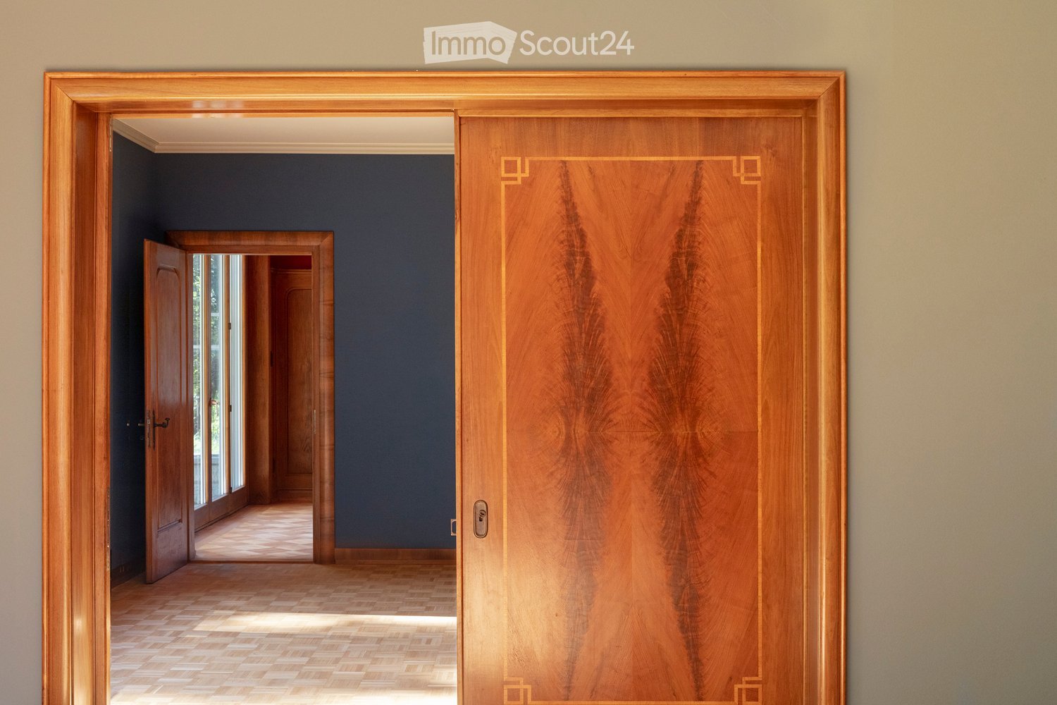 Elegant hallway with wooden doors and polished wooden flooring, leading to another room with large windows and dark blue walls.