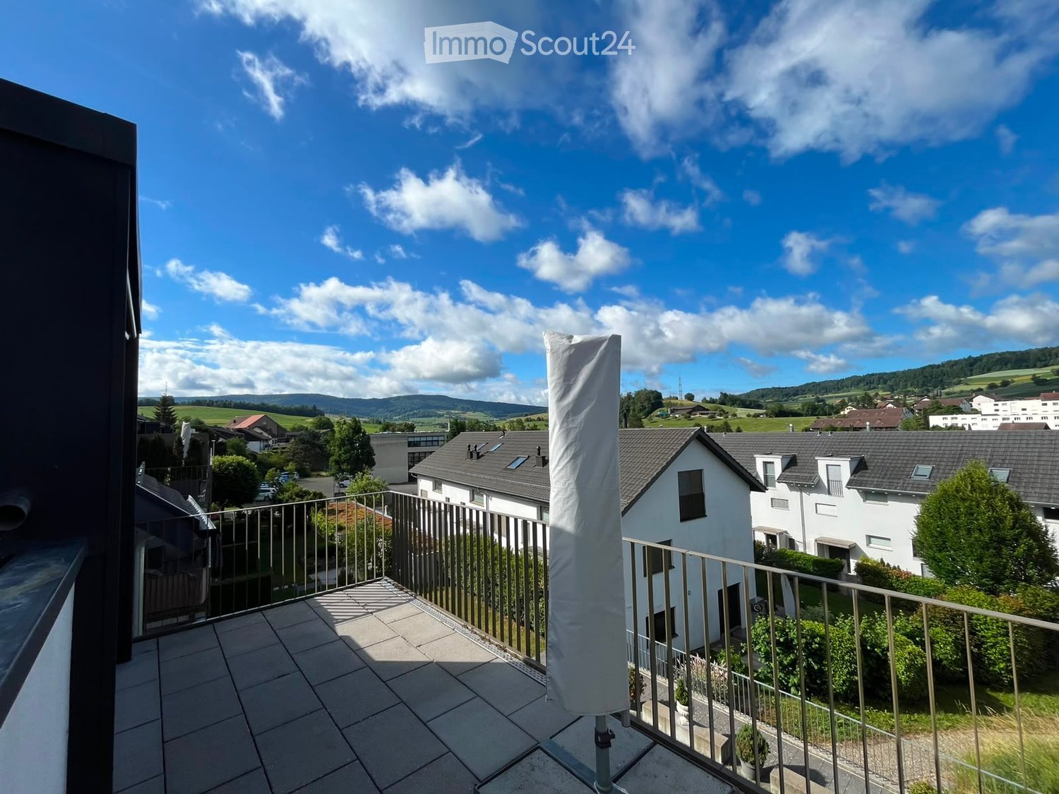 Balcony with tiled floor, metal railing, and a view of a suburban neighborhood with houses, trees, and distant hills under a partly cloudy sky.