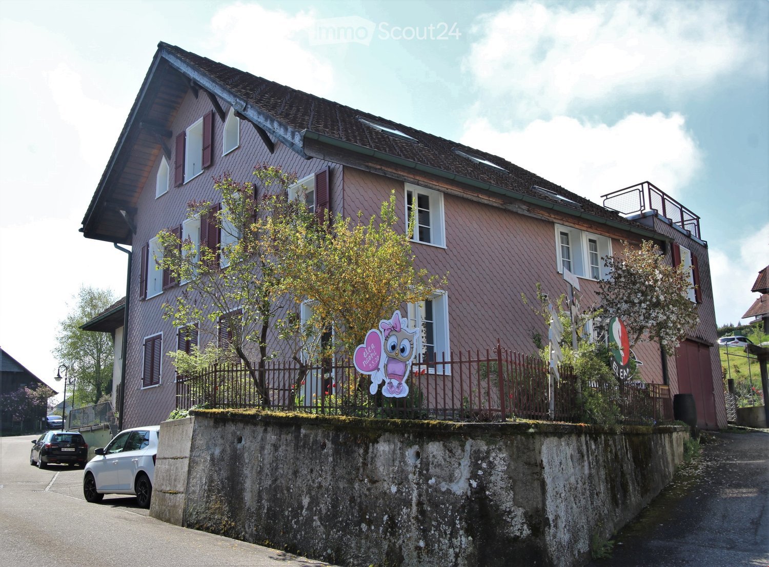 Two-story house with red-tiled roof, multiple windows, and a small balcony on the upper level. The house is surrounded by a low stone wall and a metal fence, with trees and a sign visible in front.