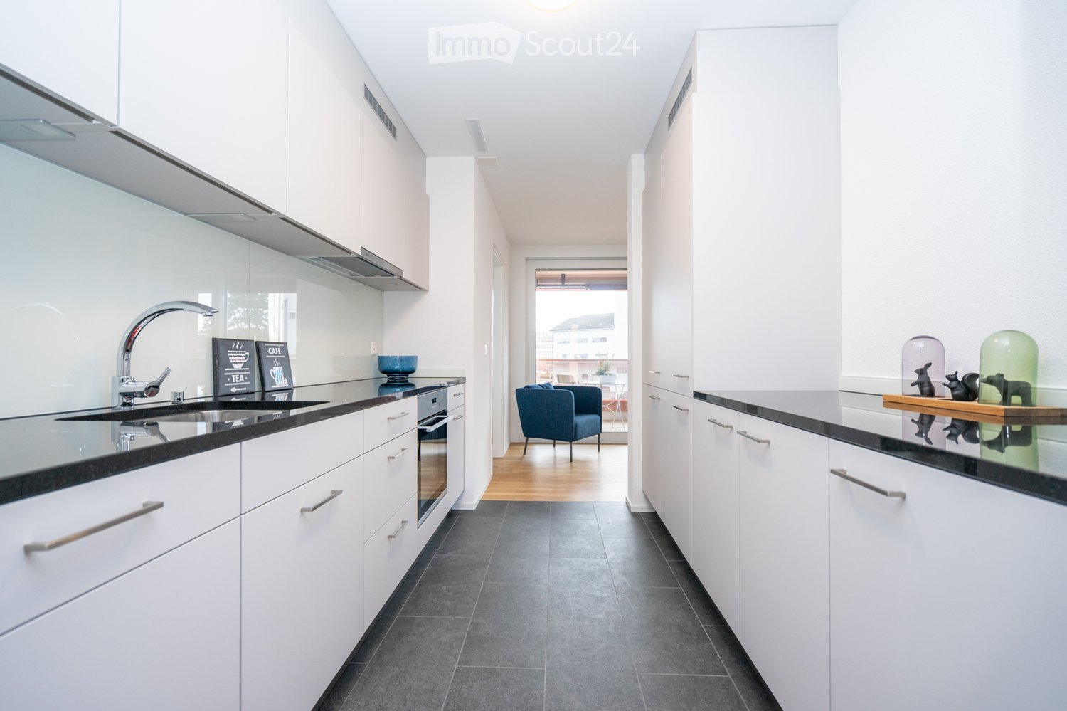 Modern kitchen with white cabinets, black countertops, stainless steel sink, and a view into a living area with a blue armchair and small table.