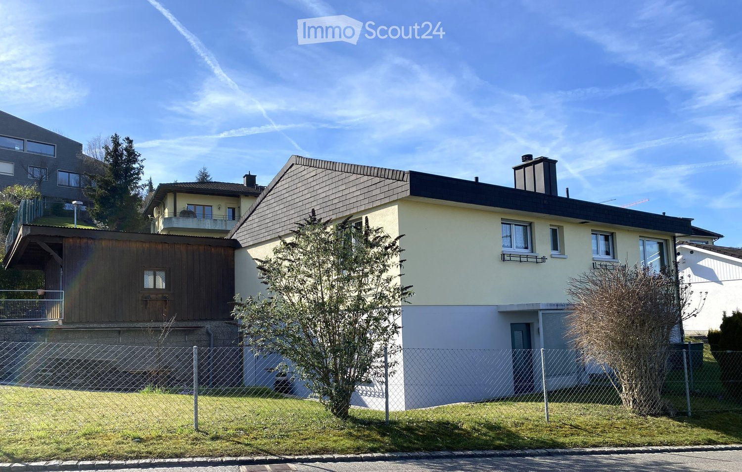 Two-story house with a combination of beige and dark brown exterior walls, multiple windows, a chimney, and a fenced front yard with trees and shrubs.