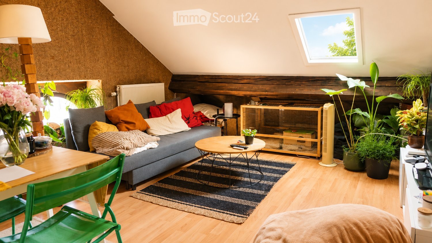 A cozy living room with a grey sofa, colorful cushions, a coffee table, a green chair, a wooden table, and several potted plants. The room features wooden flooring, a skylight, and a modern aesthetic.
