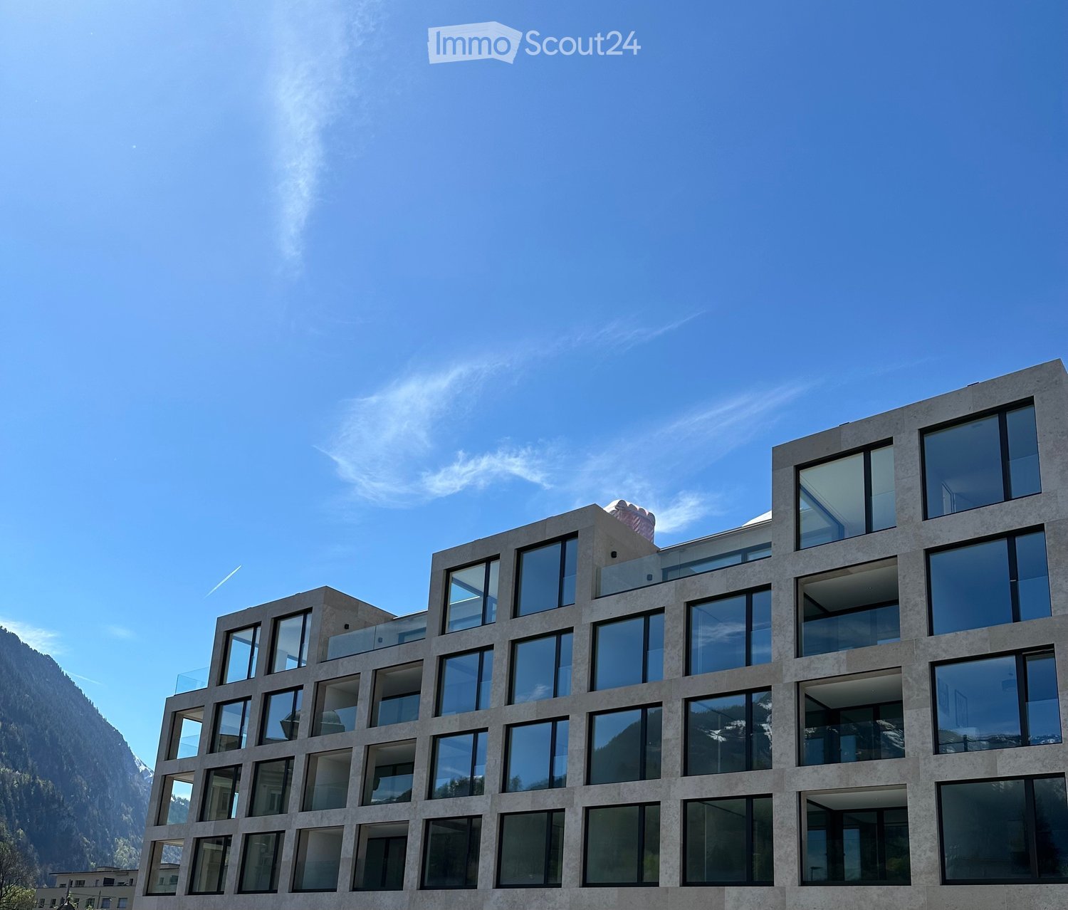 Modern multi-story building with large glass windows and balconies, set against a clear blue sky and mountain backdrop.
