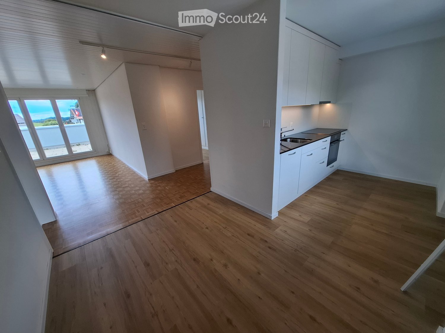 Empty room with wooden flooring, white cabinets, and a small kitchen area with sink and oven. Large window with view of outdoor area.