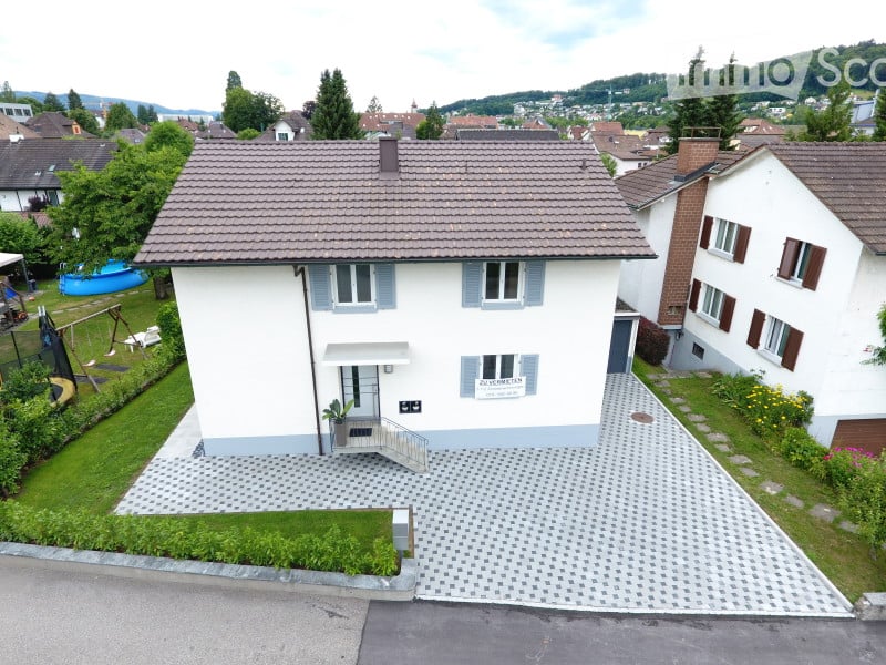 A two-story house with a tiled roof, white exterior, and blue shutters. The house features a small porch with a staircase leading to the entrance, a paved driveway, and a well-maintained garden with a swing set and a small blue pool.