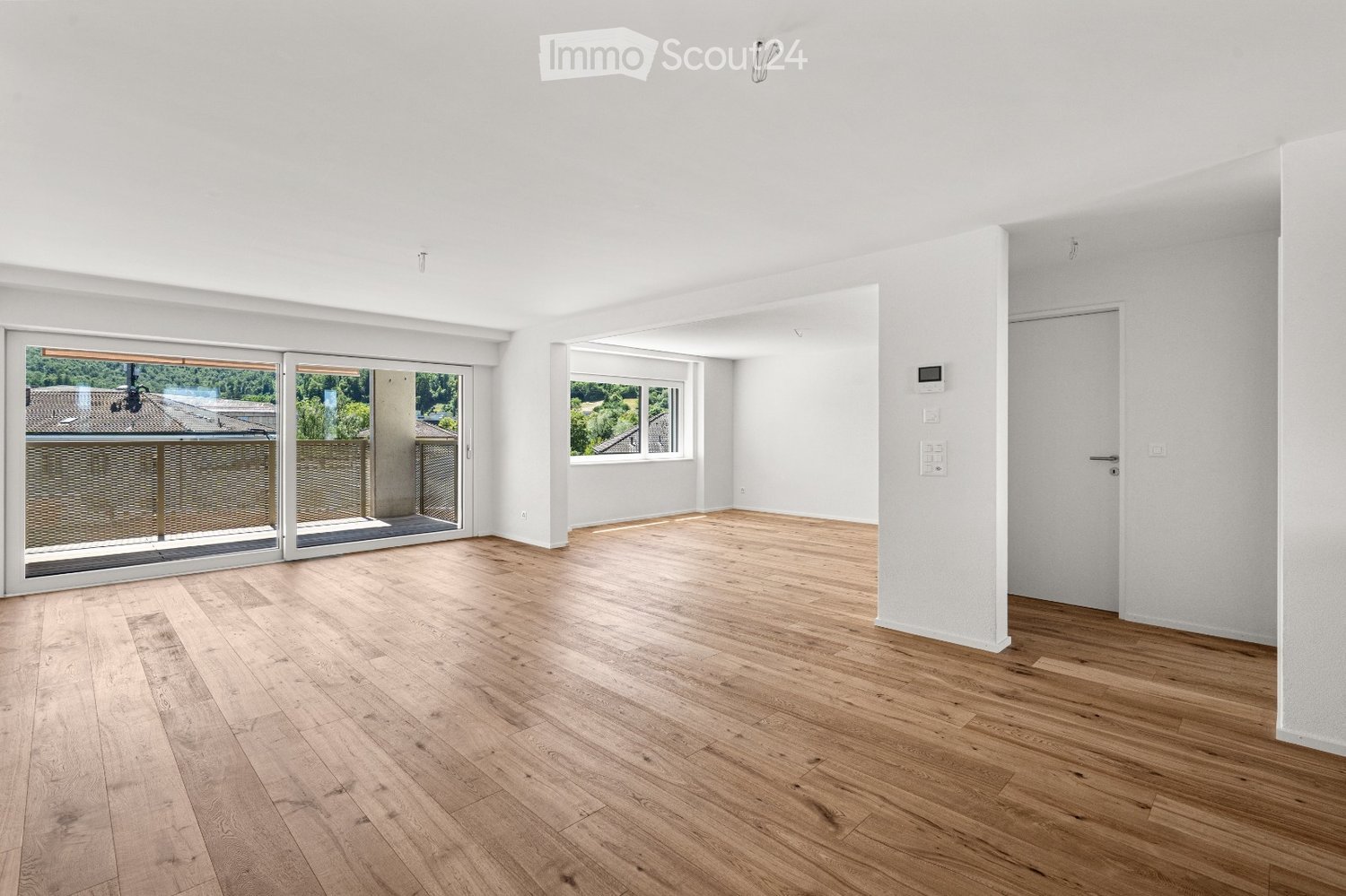 Empty room with light wooden flooring, white walls, large sliding glass doors leading to a balcony, and a window with a view of greenery.