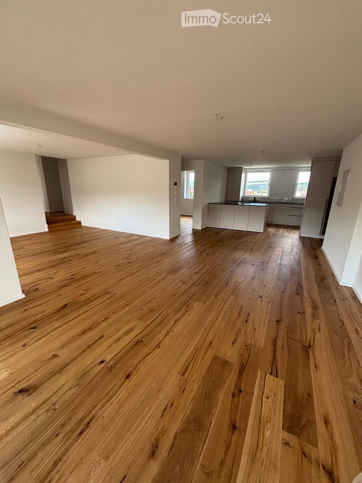 Empty room with wooden flooring and white walls, featuring an open kitchen area with cabinets and countertops visible in the background.