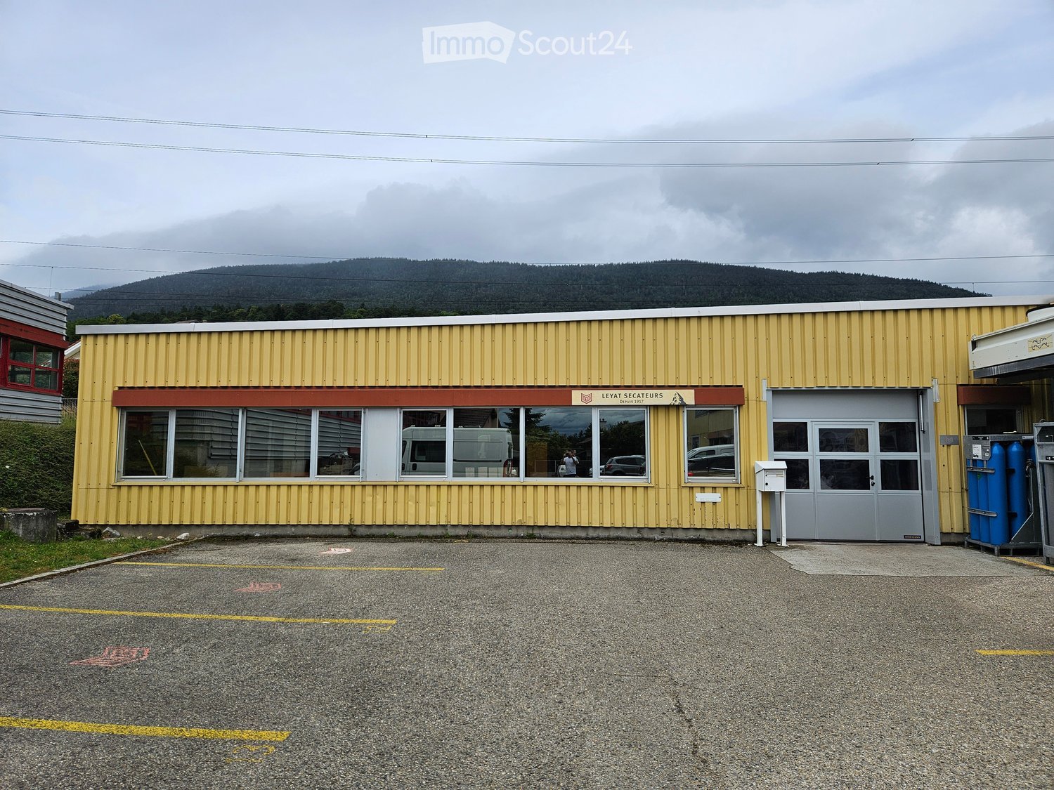 Single-story yellow building with large windows and a grey garage door, located in a parking lot with a mountain view in the background.
