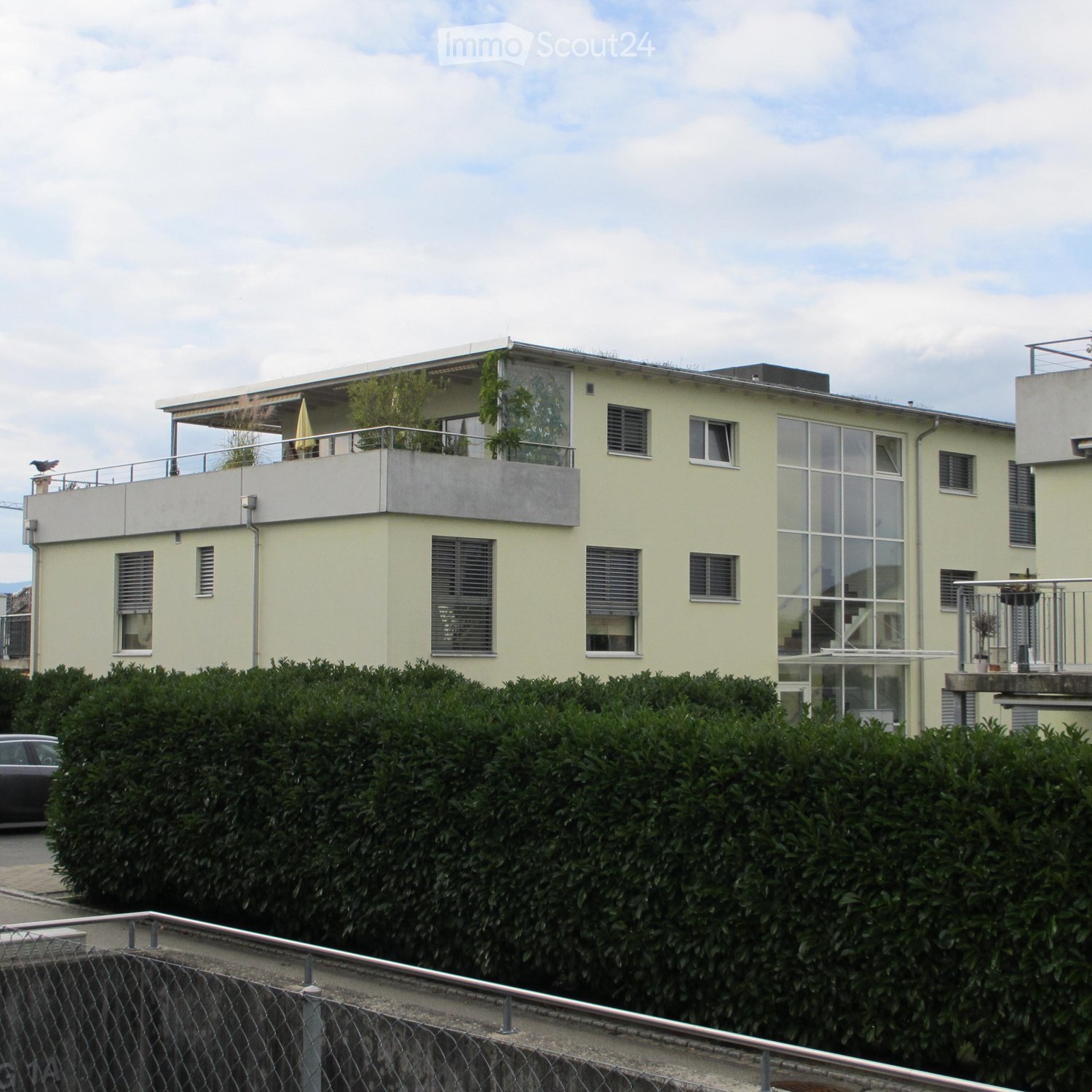 Modern two-story house with light yellow facade, large windows, and balconies. The roof features a flat terrace with plants and a railing. A hedge surrounds the property, and a car is parked nearby.
