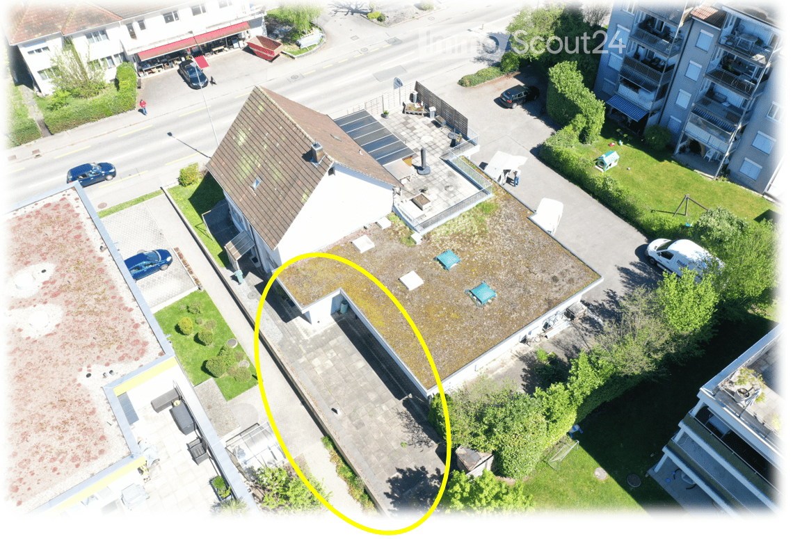 Aerial view of a multi-story residential building with a brown tiled roof, surrounded by greenery, parked cars, and neighboring structures.