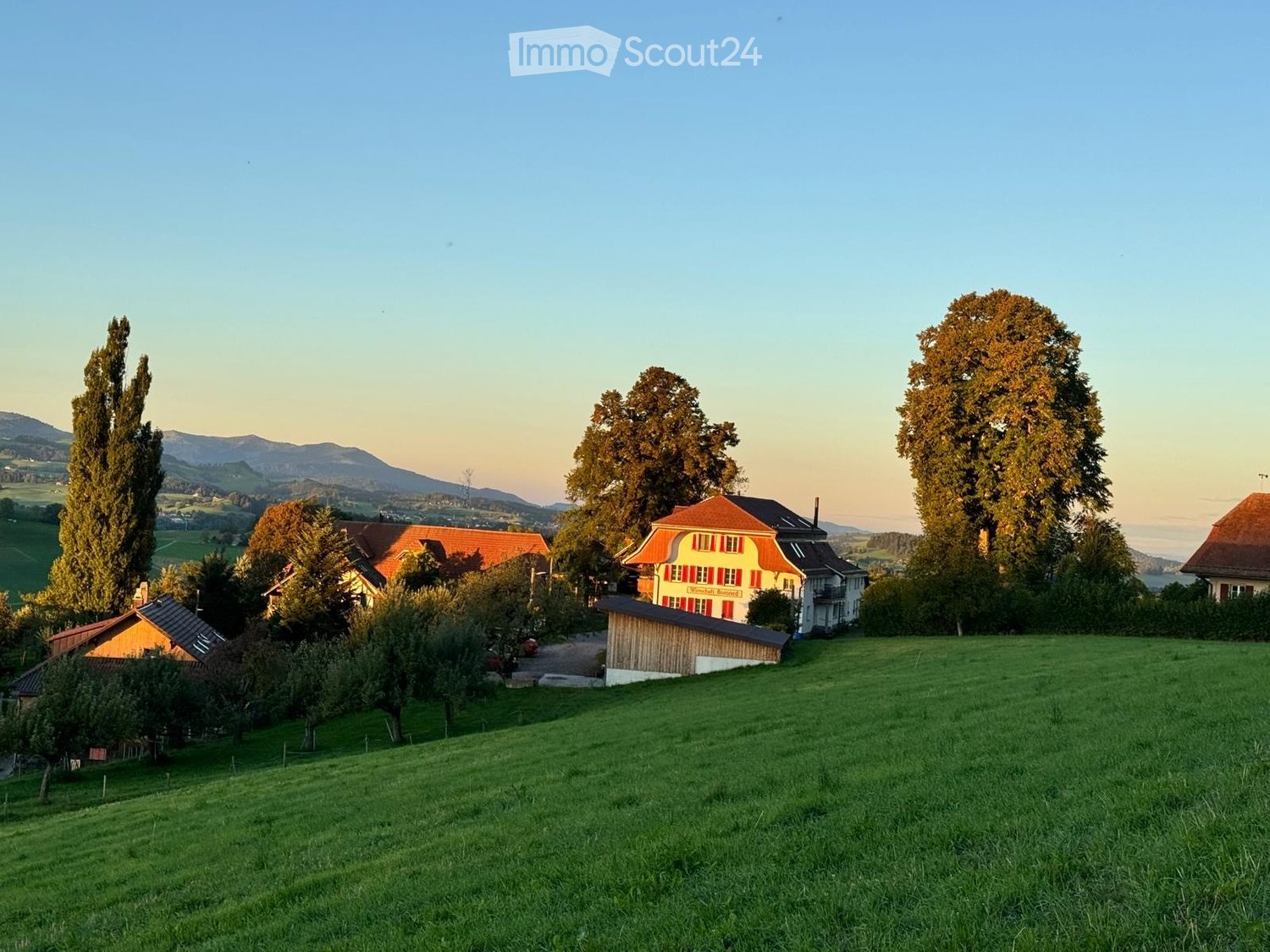 A cluster of traditional houses with red-tiled roofs and white walls, surrounded by lush green fields and tall trees, with distant mountains visible in the background.
