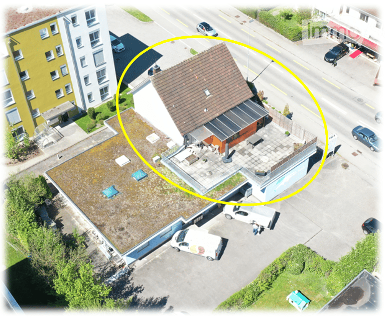 Aerial view of a multi-story building with a flat roof, surrounded by a parking lot and other structures. The building has a modern design with visible windows and a small garden area on the roof.
