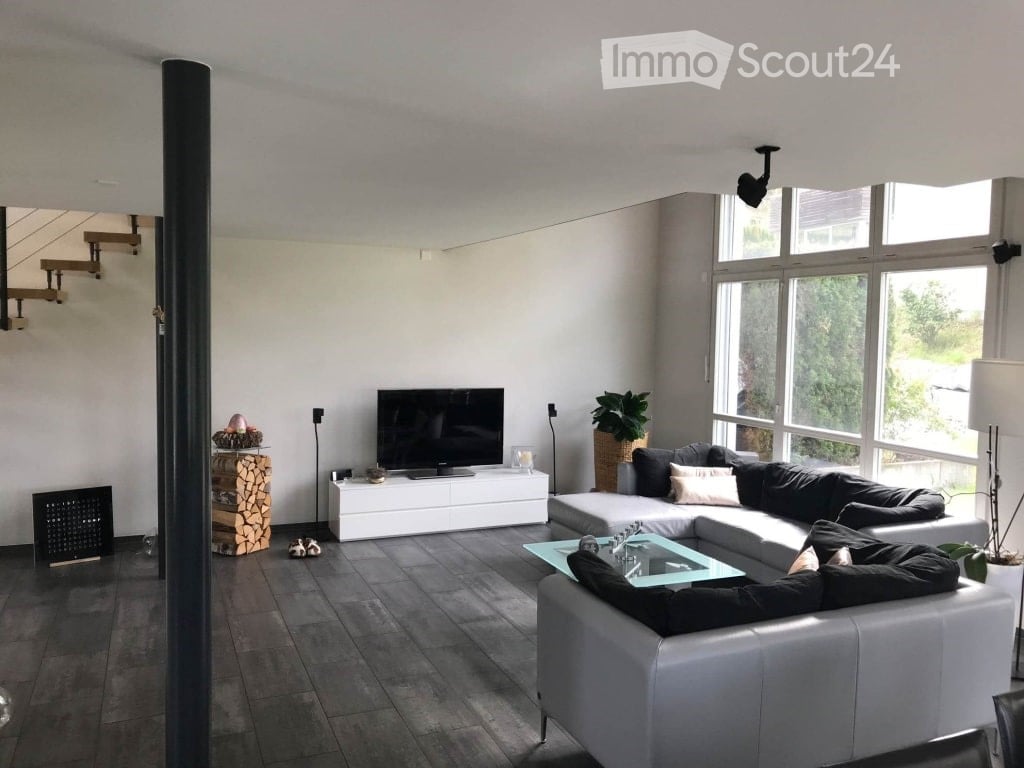 Modern living room with large windows, grey sectional sofa, glass coffee table, flat-screen TV on white stand, side table with lamp, and decorative plant in a wicker basket.